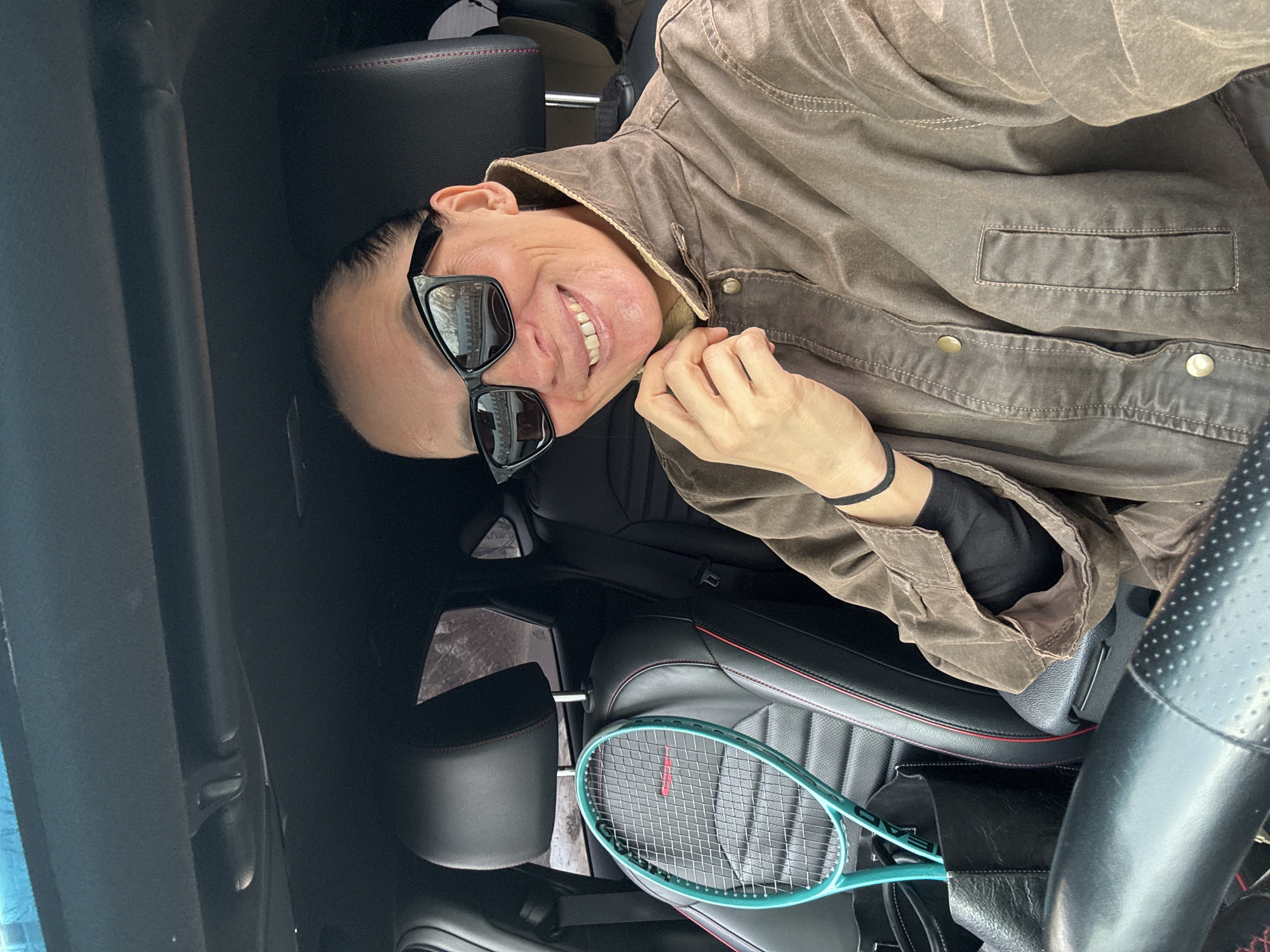 This image depicts an adult female seated in the driver's seat of a vehicle located in Chaikovsky, Russia. The female, with her dark hair pulled back, is wearing black rectangular sunglasses that reflect an outdoor scene containing bare trees and structures. She is smiling broadly, revealing her teeth, and has her left hand positioned near the collar of a brown, weathered-looking, collared jacket worn over a black long-sleeved garment. A black band is visible on her left wrist. The car's interior features black leather seats with red stitching. A turquoise tennis racket, with "HEAD" branding and a red horizontal line on its strings, is leaning against the empty front passenger seat. A black bag is also present on the passenger seat, partially obscured by the racket. A portion of the steering wheel, exhibiting a perforated grip, is visible in the lower left of the frame. The blurred background visible through the rear window shows an outdoor environment with trees or distant buildings, consistent with the reflections in the sunglasses.