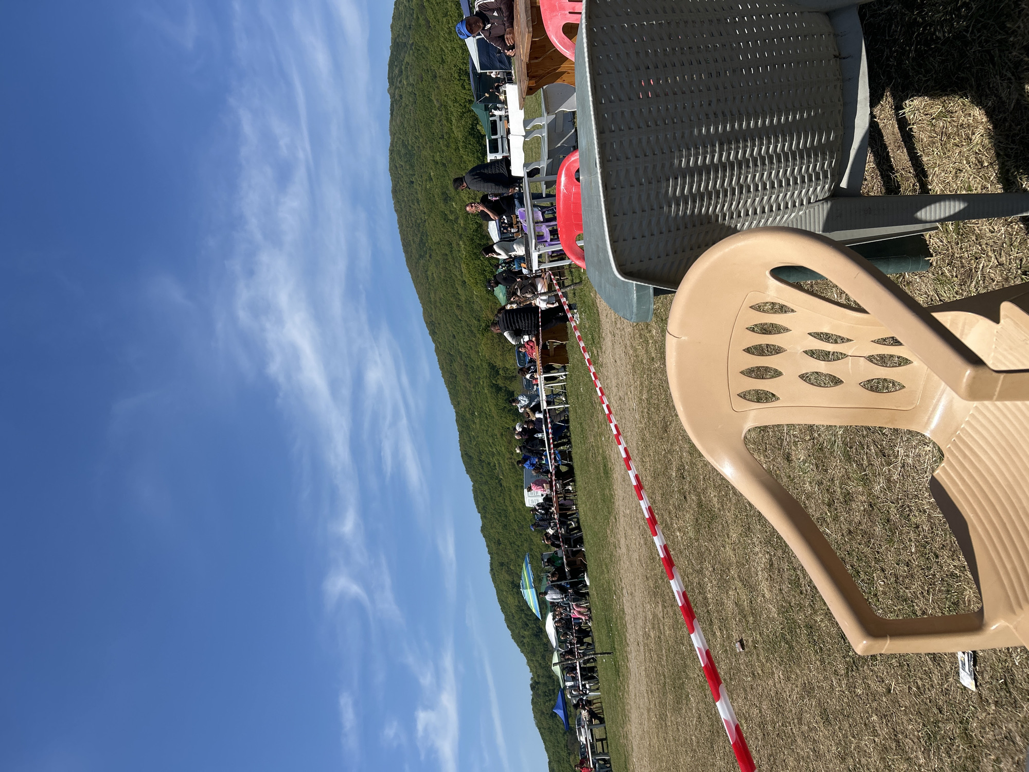 The image shows an outdoor gathering, likely an event, taking place near a grassy field with a backdrop of a green, tree-covered hill or small mountain. People are seated in rows of chairs, many of which are plastic. A red and white striped barrier runs along the length of the crowd of people. In the foreground, there are two plastic chairs, one tan and one gray. The sky is blue with some clouds. The location is likely in or near Tsarevo, Bulgaria.