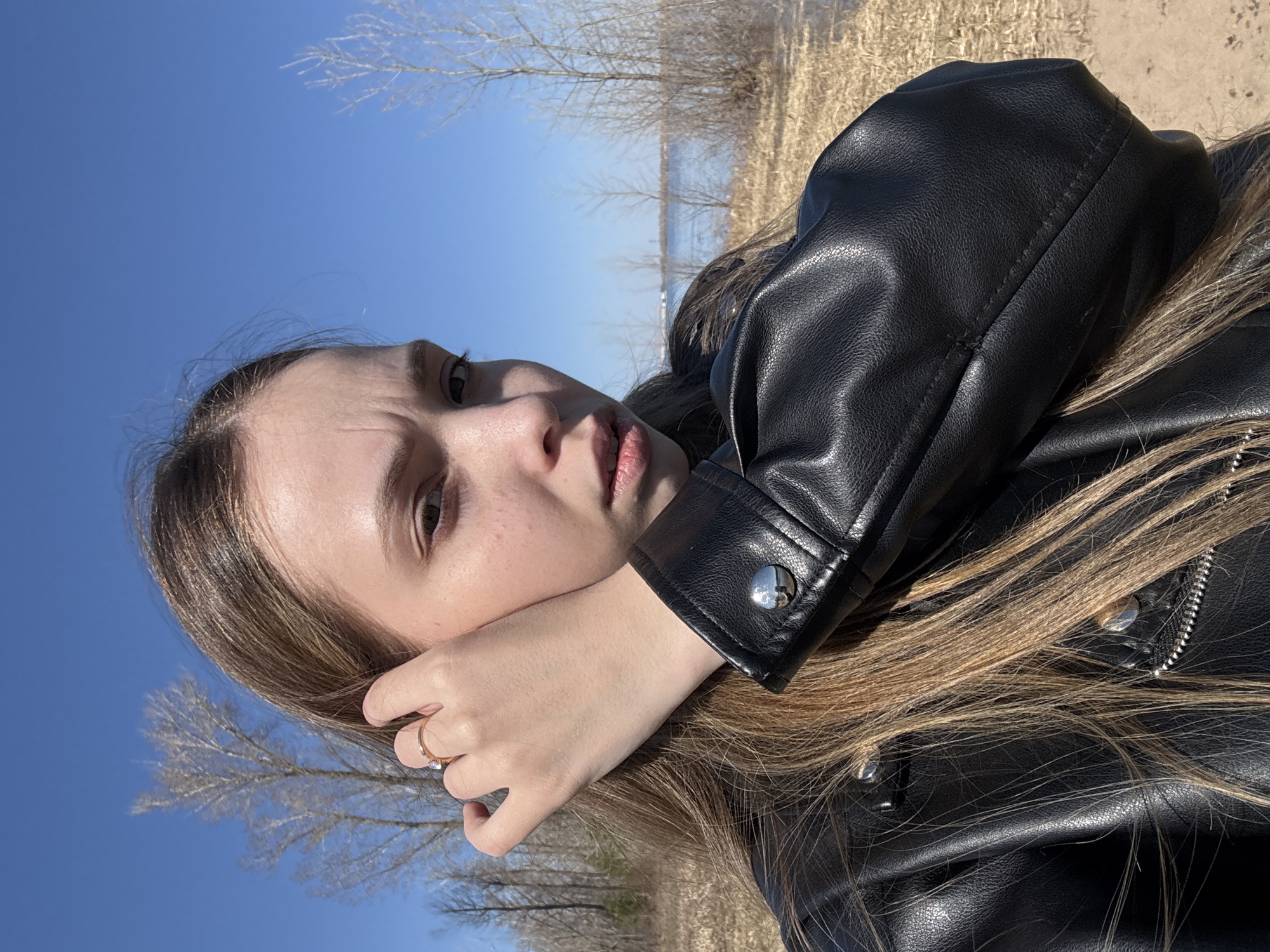 The image shows a young woman with light brown hair wearing a black leather jacket. Her face is partially obscured by her hand, on which she wears a ring. The background suggests an outdoor setting near a body of water, with trees visible against a blue sky. The location is Прикамский, Russia.