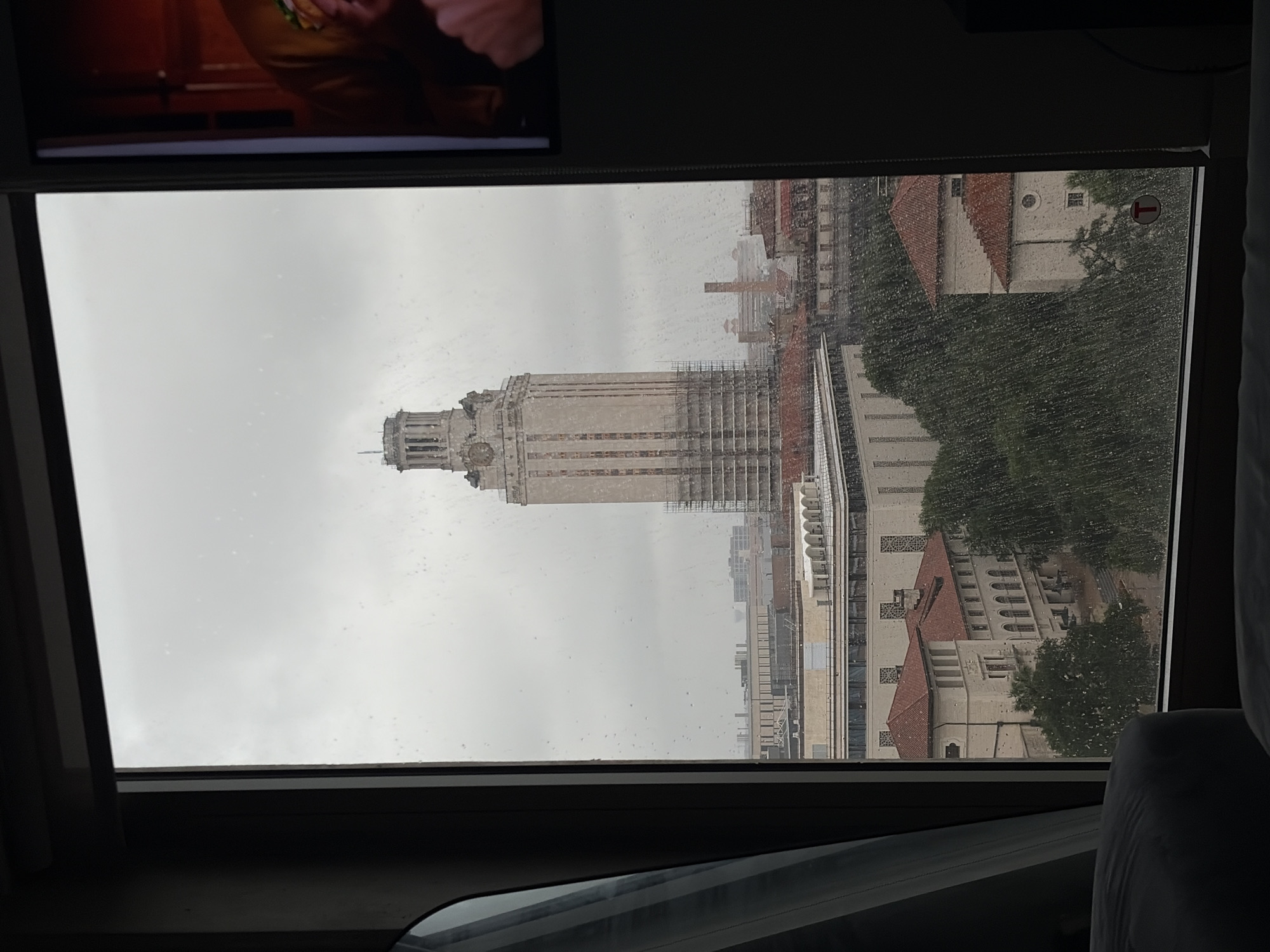 The image captures a view through a rain-splattered window, looking out at the University of Texas at Austin campus on an overcast day. The central subject is the iconic UT Tower, a tall, light-colored stone structure, which has extensive scaffolding on its right side, indicating construction or maintenance work. Surrounding the tower are other university buildings with traditional red-tiled roofs and intricate architectural details, partially visible amidst green trees. The sky is uniformly grey and cloudy, contributing to the subdued lighting, and numerous raindrops are clearly visible on the windowpane, suggesting active rainfall or recent rain.
In the upper section of the image, the frame of the window is visible, and above it, a reflection or a display screen shows a blurry, warm-toned image of a person's upper body. The lower right portion of the image shows a dark, upholstered surface, possibly a headrest or seat interior, suggesting the photo was taken from inside a vehicle or a room with an unconventional window placement. A small, circular red sign with a white arrow pointing to the right is affixed to the window on the far right side. The scene conveys a quiet, possibly damp, daytime atmosphere on the university campus.