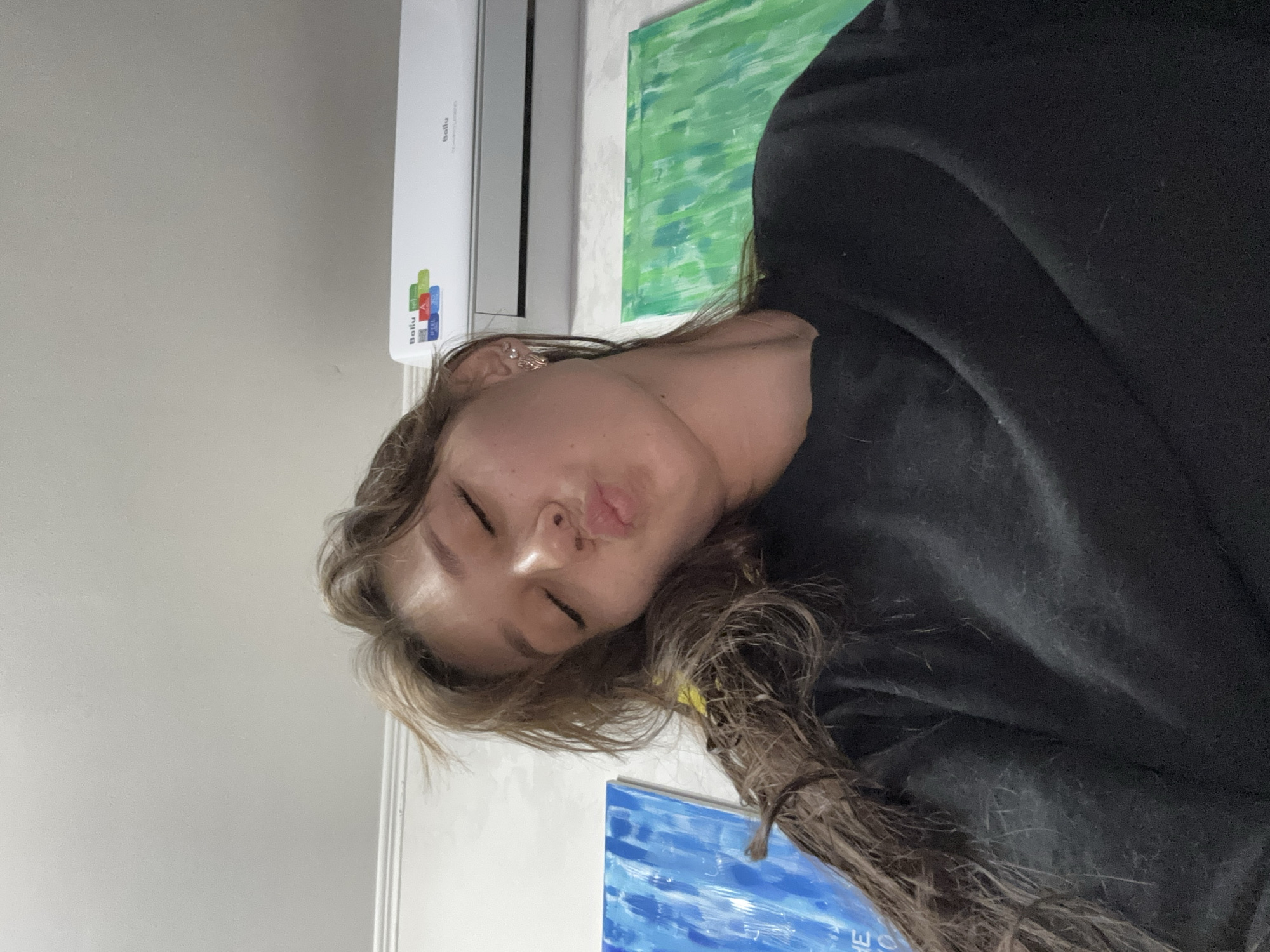 The image shows a young woman with her eyes closed and lips pursed, as if blowing a kiss. She has long, light brown hair that is falling over her left shoulder. She is wearing a black t-shirt. Behind her, an air conditioner is mounted on the wall. To her left, there is a painting with blue and white vertical brushstrokes. To her right, there is a painting with green and white vertical brushstrokes. The overall environment appears to be a room in a residential building. There is no indication of the specific location within Novokuznetsk, Russia.