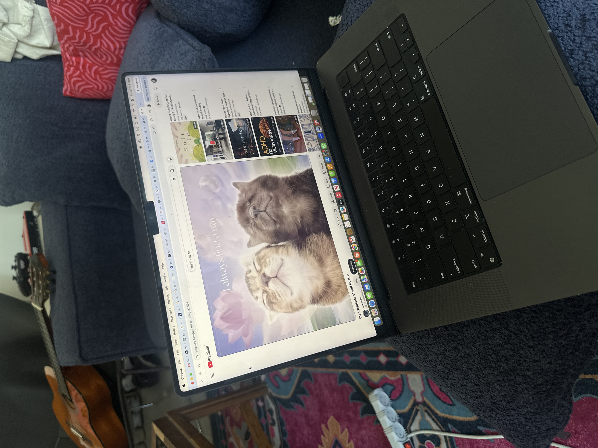 The image shows a person's lap and legs covered by a dark blue textured blanket, with a laptop open on their lap. The laptop displays a YouTube video. The video appears to be a relaxation or meditation video, with an image of two cats and the text "I always find a way." The laptop is a MacBook Pro, identifiable by its design and the Apple logo. The keyboard and trackpad are visible.
In the background, a dark blue couch is visible, with a guitar resting on it. There are also some pillows on the couch, one with a red abstract pattern. A colorful rug is visible on the floor, and an electrical power strip with several plugs is partially visible on the left side of the image.
The image was taken indoors, likely in a living room. The lighting appears to be natural, possibly from a window. There is no indication of the specific location in Tel Aviv, Israel.