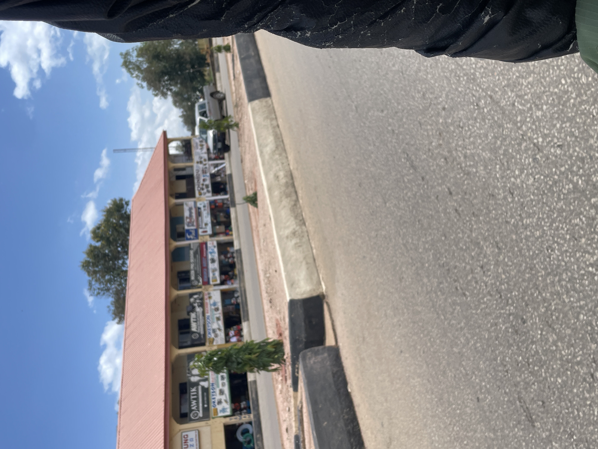 This is a vertical, eye-level, medium shot of a street scene in Gura Top, Nigeria. The image is dominated by a road, a sidewalk, and a building with multiple shops.
The road is paved and appears to be a bit worn, with speckled grey asphalt. To the left of the road is a concrete sidewalk with a curb. A black and grey concrete barrier runs along the edge of the sidewalk.
On the other side of the sidewalk is a multi-story building with a light pink, sloped roof. The building has several shopfronts on the ground floor, some of which are visible. Signs are displayed outside these shops. One prominent sign reads "AWTİK". Another partially visible sign reads "OKEYSON". Another sign has a logo with the letters "UNQ" and what appears to be "ZO". There are people visible behind the shop windows, suggesting the shops are open.
Behind the building, trees are visible, and the sky is a bright blue with scattered white clouds. The sunlight suggests it is daytime, likely mid-morning or afternoon. A white vehicle is parked on the side of the road near the building.
The image is taken from a slightly elevated perspective, possibly from a moving vehicle or a person standing. The overall mood is neutral, depicting an everyday street scene.