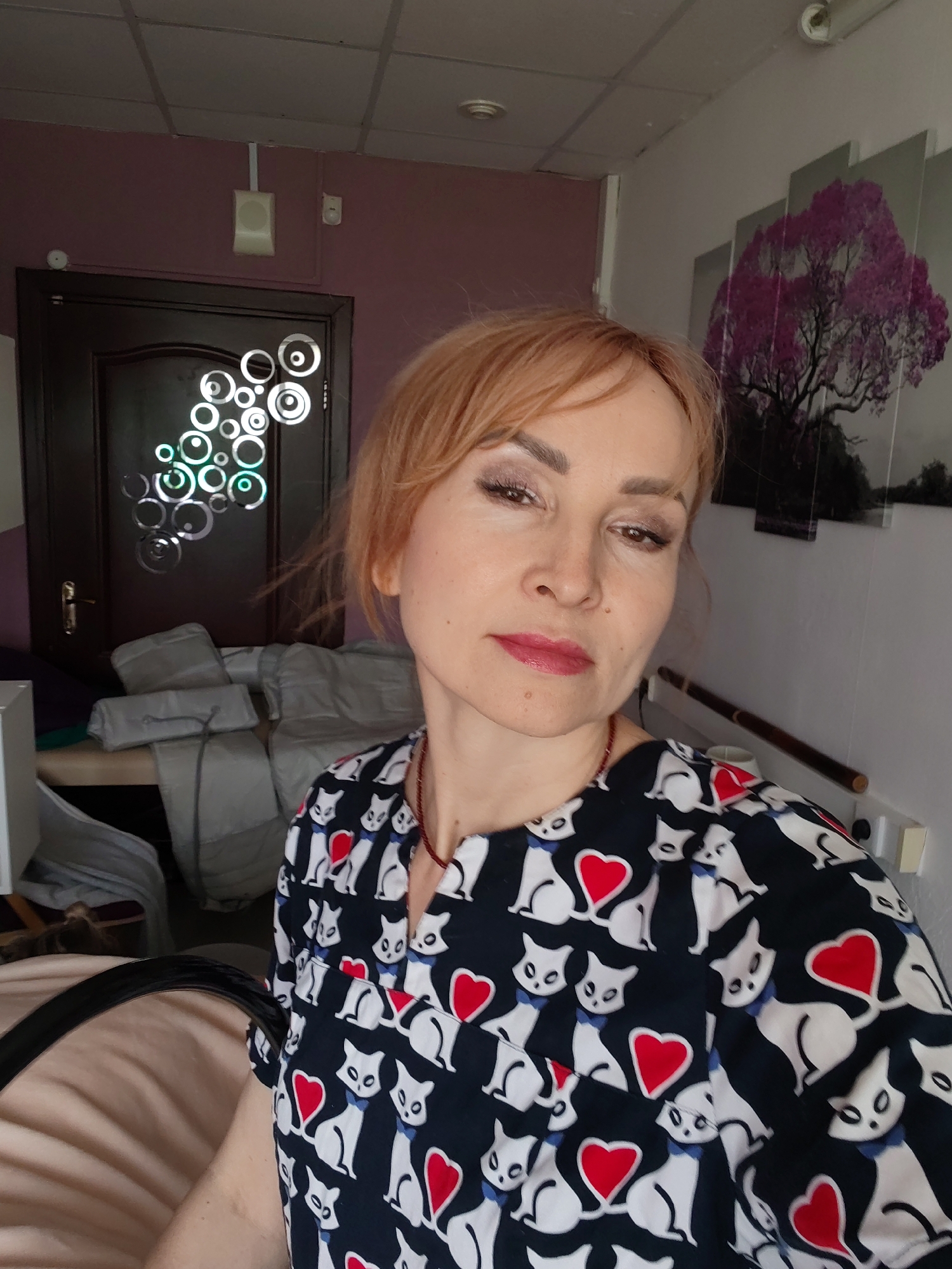 The image shows a middle-aged woman posing for a selfie. She has light red hair and is wearing a dark blue top with a pattern of white cats and red hearts. She has lipstick on.
Behind her is a door with decorative white circles. To the right of the door, the wall is purple, and further to the right, the wall is white and decorated with a multi-panel artwork depicting a tree. A medical or treatment table covered in gray linens is visible to the left. A handrail and a light switch are visible on the white wall.
While the user prompts states the location is in the city of Chaikovsky, Russia, there is nothing within the image itself that indicates the geographical location. Therefore, location information will not be added.