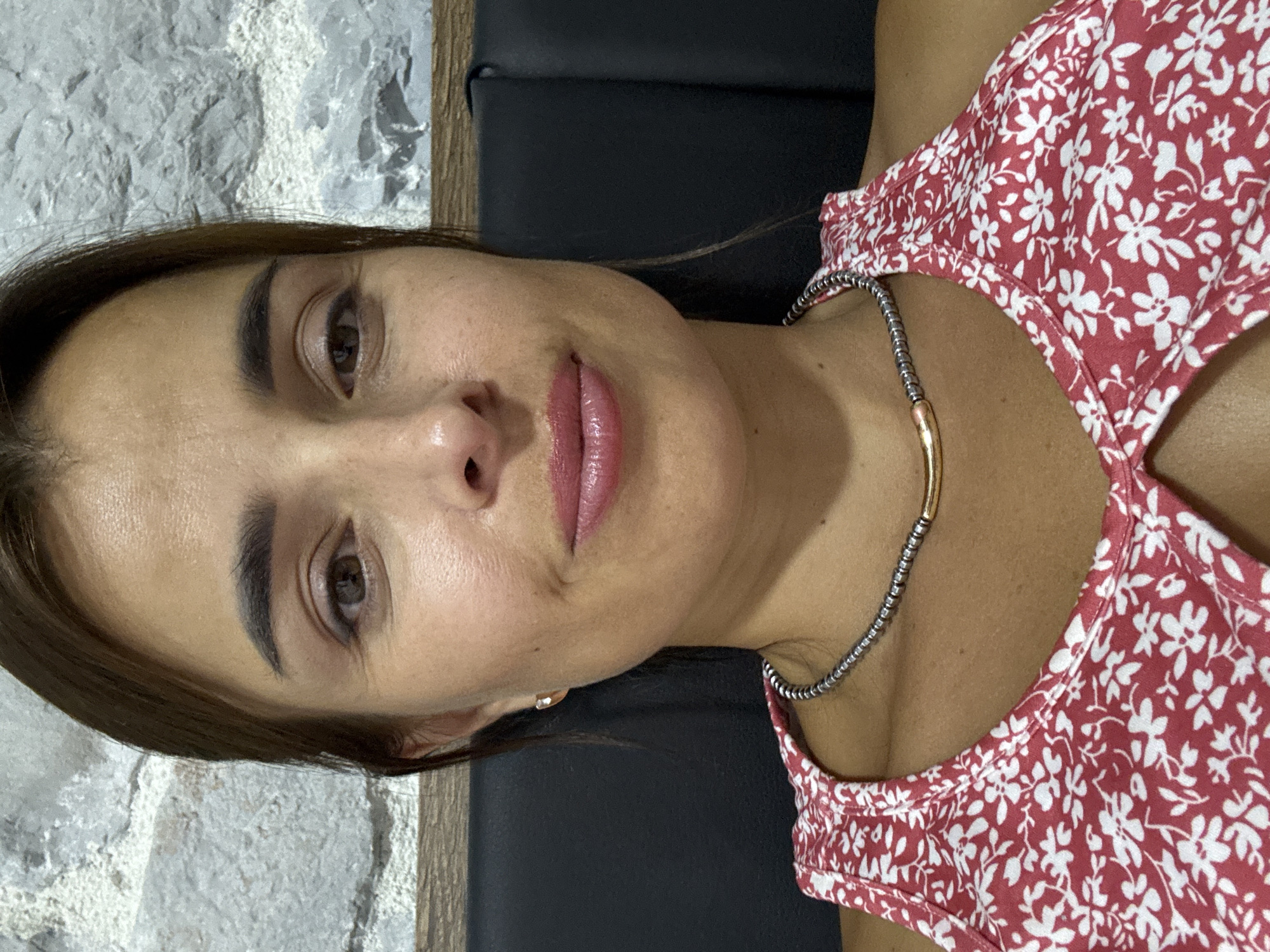 A woman is pictured in a close-up, head and shoulders shot. She has brown hair pulled back, brown eyes, and is wearing a red and white floral top. She also has a necklace with silver beads and a gold accent. The background appears to be a textured stone wall and a dark upholstered surface. The setting is indoors.