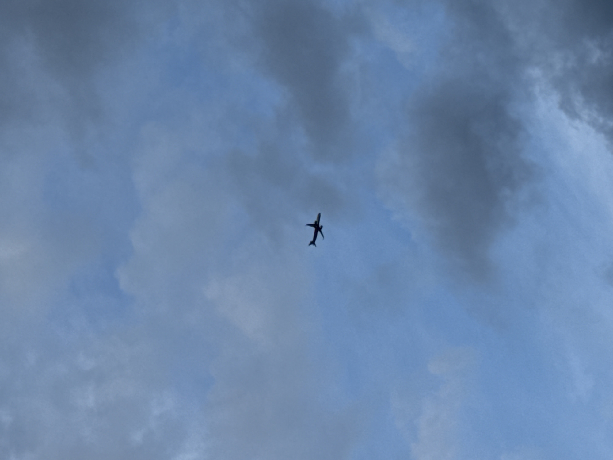 A dark silhouette of a passenger airplane is captured in mid-flight against a cloudy blue sky. The aircraft is positioned slightly left of the vertical center of the frame, with its nose pointing towards the upper left. The clouds in the background are a mix of light and dark gray, indicating overcast or partially cloudy conditions. The airplane appears to be a commercial jet with visible wings and tail. The scene is devoid of any ground-level context, suggesting the photograph was taken from a location with a clear view of the sky.