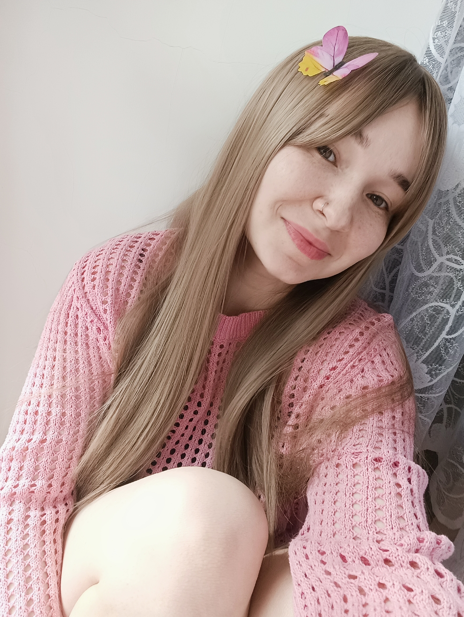 A young woman with fair skin, light brown hair with bangs, and freckles is posing for a close-up selfie. She is wearing a pink knitted sweater and has a butterfly hair clip on the right side of her head. She is smiling and has a nose ring. The background consists of a white wall on the left and a white lace curtain on the right.
There is no indication in the image that this was taken in the city of Izhevsk, Russia.