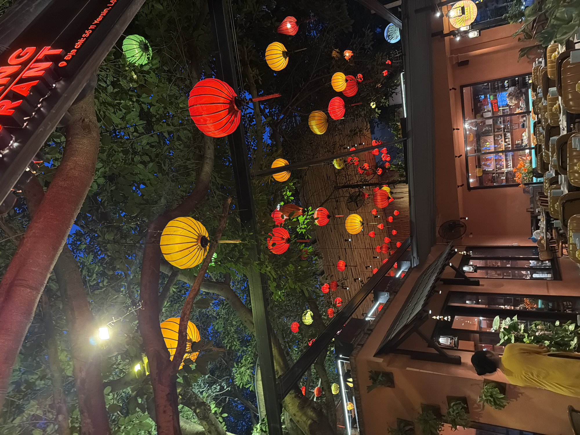 The image depicts an outdoor dining area at a restaurant in Nha Trang Ward, Vietnam, at dusk. The foreground shows tables and chairs set up for patrons. A woman in a yellow dress stands with her back to the camera, looking towards the building's entrance. The building is painted an orange hue, with large glass doors and windows revealing shelves filled with various items inside. Hanging lanterns in red, yellow, and green are strung throughout the trees and overhead canopy, creating a festive atmosphere. The branches of large trees with lush green foliage extend over the dining area. In the background, a neon sign that reads "NG RANT" is visible, suggesting the establishment's name. The overall scene suggests a vibrant and inviting dining experience in a tropical setting.