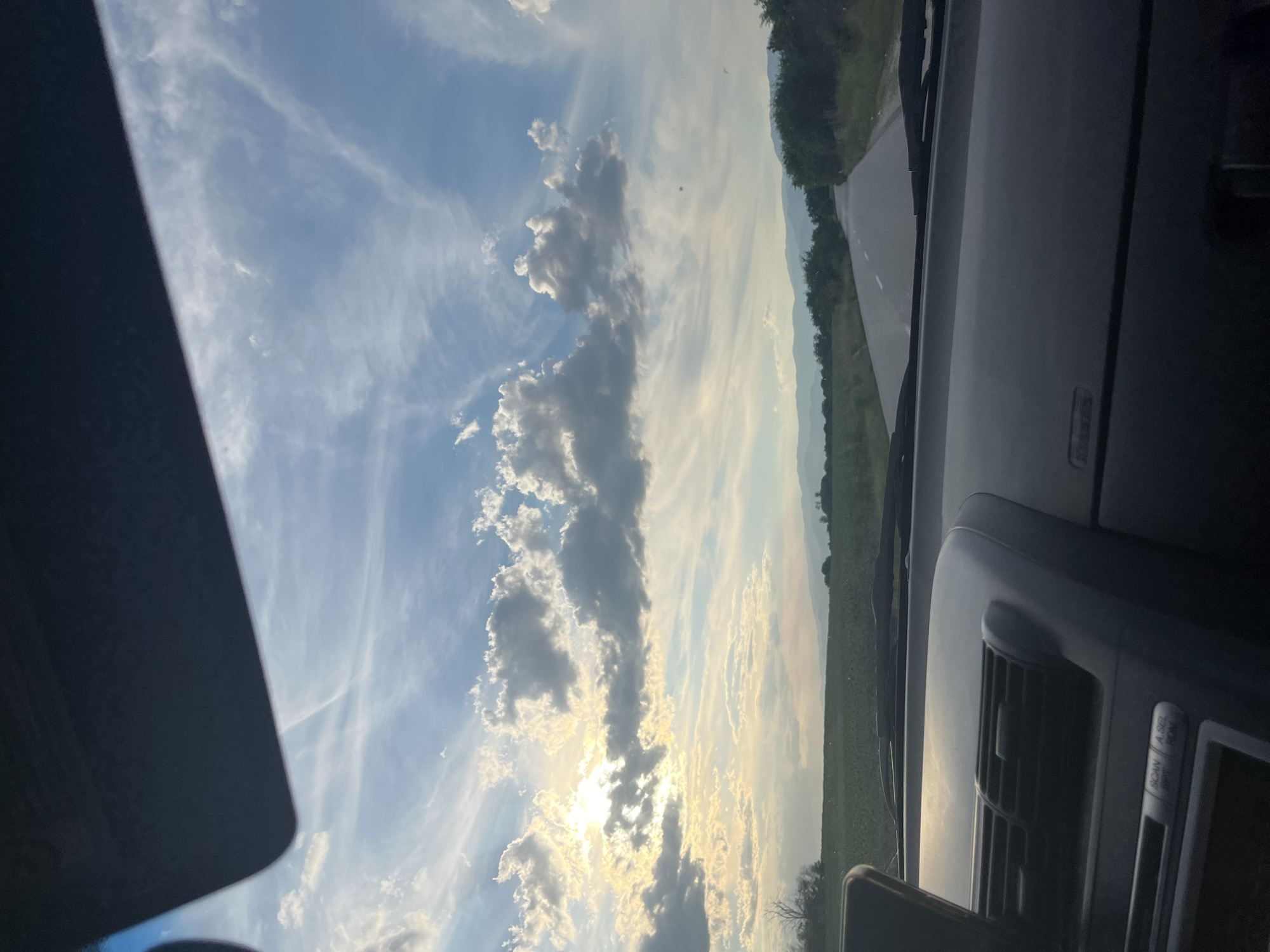 The image, taken from inside a vehicle, captures a view of a rural road in Zhitnitsa, Bulgaria, under a sky filled with dramatic clouds and the setting sun. The foreground is dominated by the dashboard of the car, featuring an air vent and part of the car's audio system. A dark sun visor is visible at the top of the frame.
The midground shows a paved road stretching into the distance, bordered by green fields on the left and trees and vegetation on the right. The fields appear cultivated, with some dark elements suggesting agricultural activity or equipment. In the far distance, a range of hills or mountains is visible, softened by the atmospheric haze.
The sky is a prominent feature, with a mix of bright sunlight breaking through scattered clouds and wispy cirrus clouds that create a streaky effect across the blue expanse. The sun itself is partially obscured by clouds, casting a warm, golden light that illuminates the scene. The overall impression is one of a peaceful, end-of-day drive through the Bulgarian countryside. There are no people or animals visible in the image.