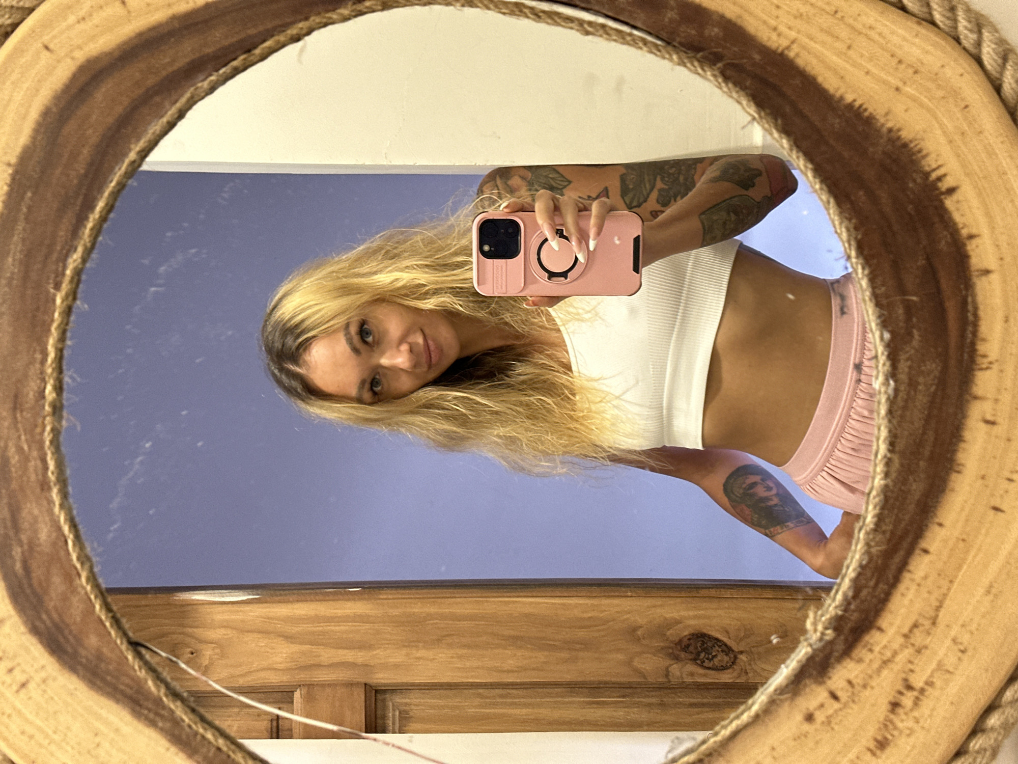 This is a mirror selfie taken by a woman with long blonde hair and tattoos on her arms. She is wearing a white crop top and pink shorts. She is holding a pink phone in her right hand, using it to take the photo. The background is a wall painted in a light purple color, and there is a wooden frame around the mirror. The lighting appears to be indoor, and the mood is casual and relaxed. The photo is taken from a slightly angled perspective, showing the woman's face and upper body.