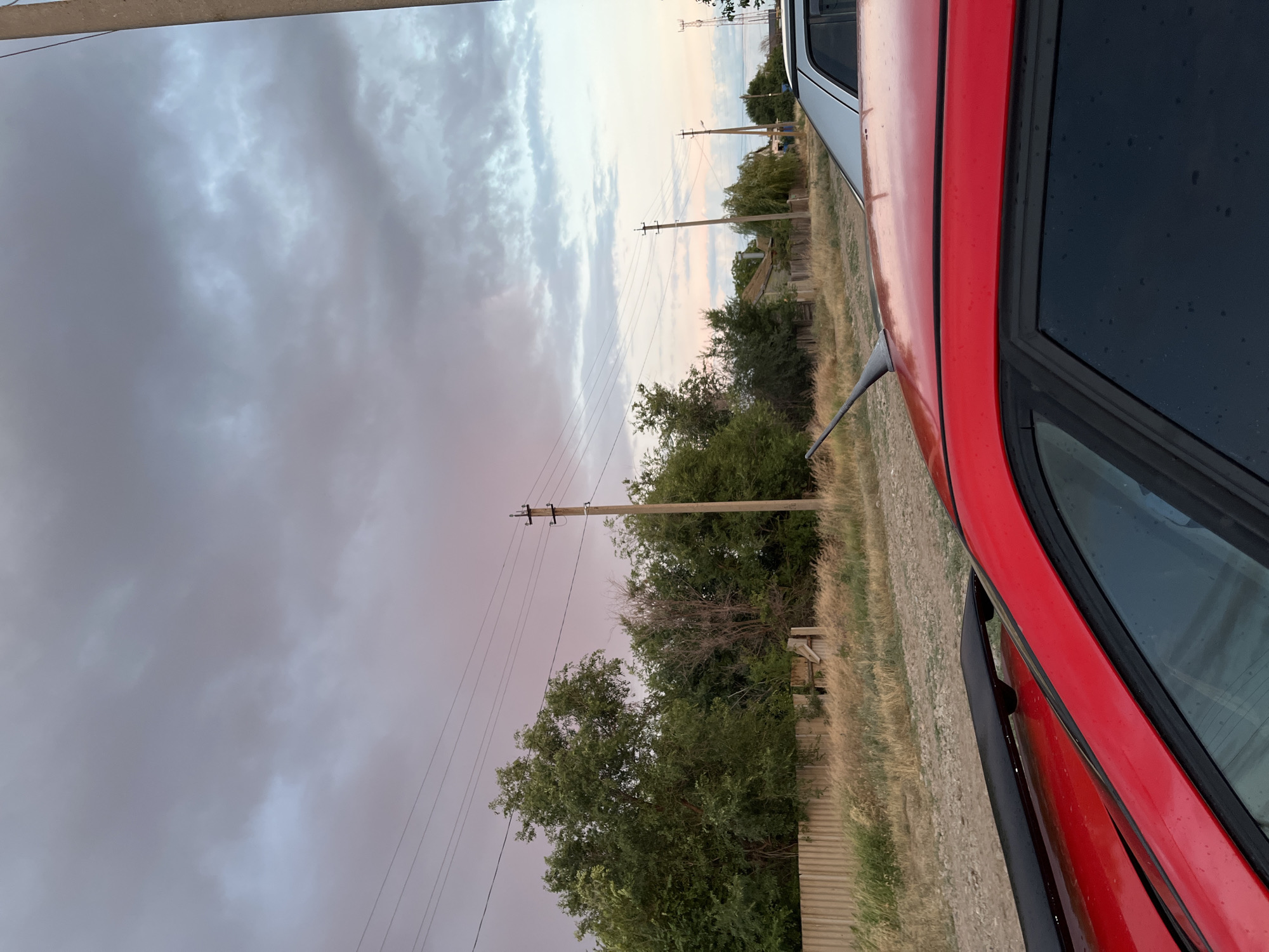 The image shows a rural street scene in Rakhinka, Russia, with a dramatic sky. In the foreground, the top and side of a red car are visible, its surface showing water droplets. Behind the car, dry, tall grass lines a dirt road. A wooden fence runs along the edge of the property, with dense green trees and some dry brush behind it. Several utility poles with power lines crisscross the sky, which is filled with heavy, gray clouds tinged with pink and pale blue from the setting sun. In the middle ground, houses with wooden fences are visible amongst the trees. Another car, silver or white, is parked further down the road. The overall impression is one of a quiet, possibly impending storm in a rural, residential area.