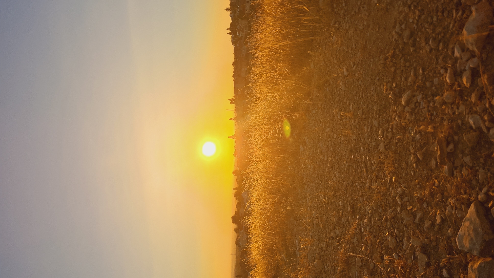 The image shows a landscape at sunset in Be’er-Sheva, Israel. The sun is positioned low on the horizon, radiating a golden light across the scene. The sky transitions from a pale blue to orange as it nears the sunset point. The foreground consists of a rocky, uneven ground surface interspersed with dry grass. In the distance, there is a sparse residential area, with a few buildings visible against the horizon. There are no people visible in the scene.