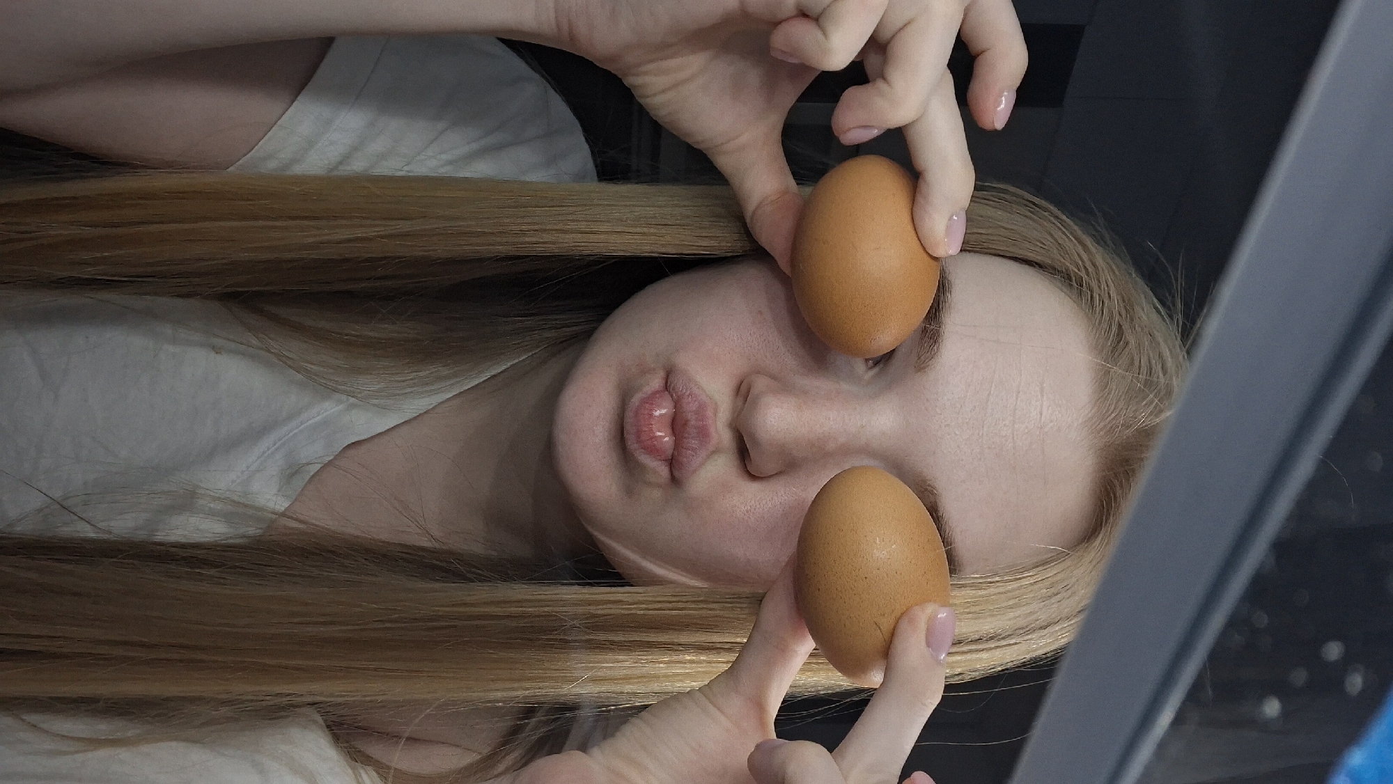 A woman with long, blonde hair is holding two brown eggs in front of her eyes. Her lips are puckered. The background is a dark gray or black surface, possibly a wall or appliance. The location of the image is presumed to be in Chaikovsky, Russia.