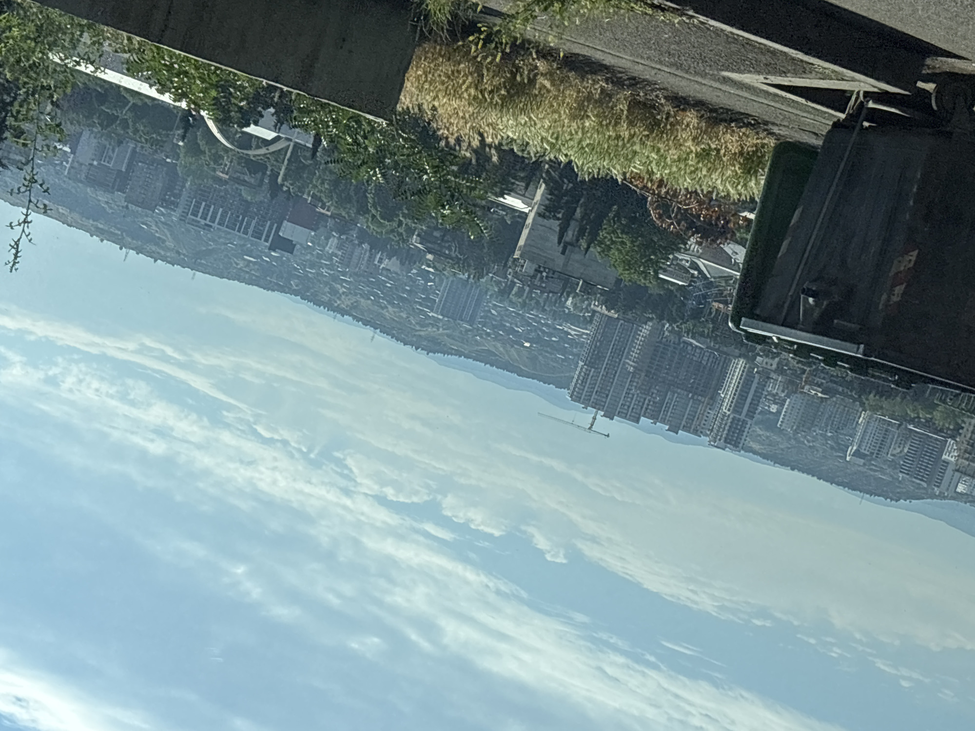 The image depicts a panoramic view of Tbilisi, Georgia, from an elevated vantage point. In the foreground, a large, dark green dumpster is positioned on the left side of the frame. It is angled towards the right, with visible wheels and a lid. Adjacent to the dumpster, a concrete barrier runs horizontally across the lower portion of the image. Beyond these elements, the landscape unfolds to reveal a sprawling cityscape. Numerous apartment buildings, varying in height and architectural style, are densely packed together on a hilly terrain. Some buildings are modern, with numerous floors and balconies, while others appear older and lower in profile. A construction crane is visible in the mid-ground, suggesting ongoing development. Lush green trees and vegetation are interspersed throughout the urban landscape, particularly on the slopes of the hills. The sky is a clear, bright blue, adorned with wispy white clouds that stretch across the horizon. The overall scene suggests a day with fair weather.
