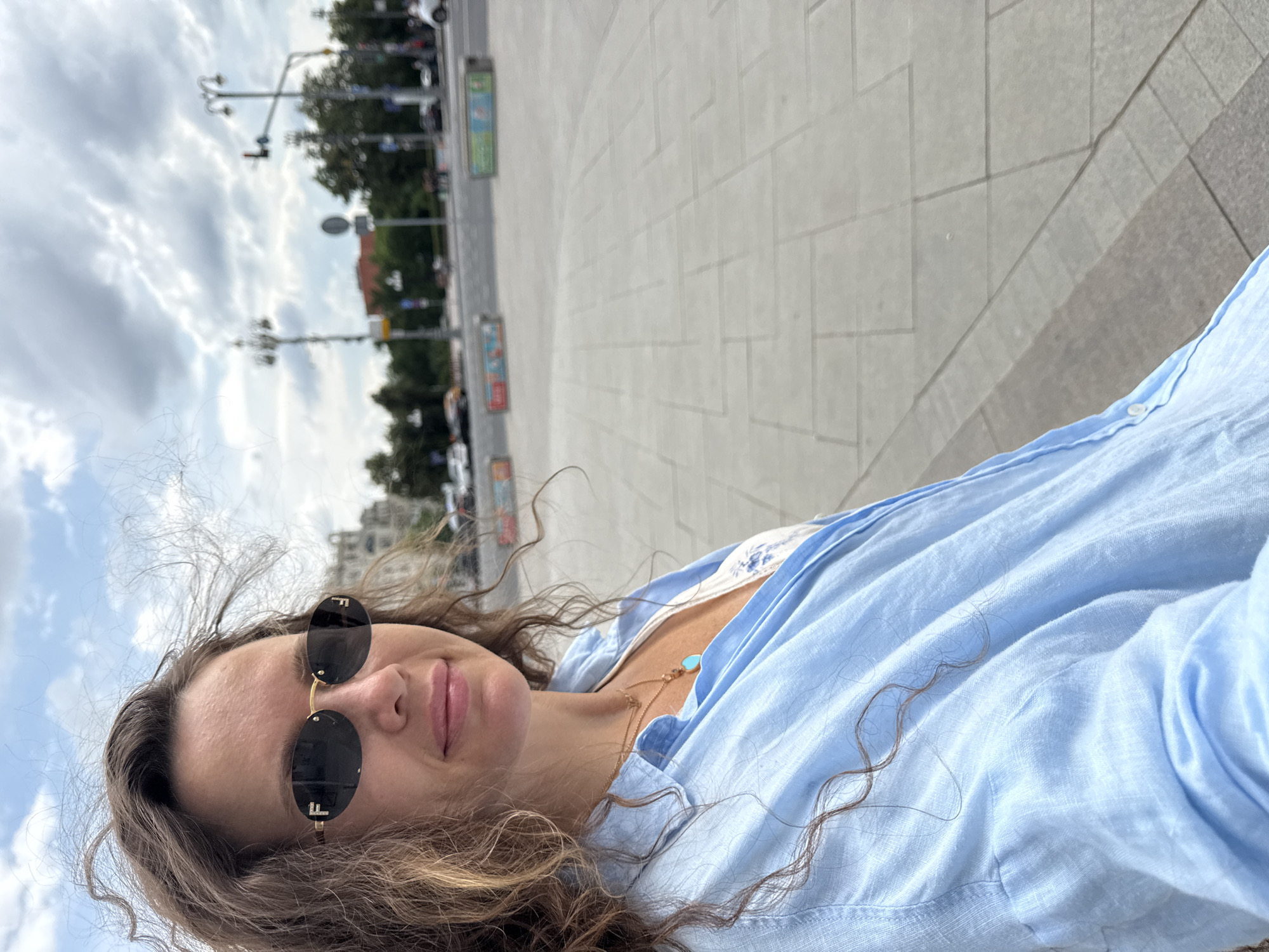 A woman wearing sunglasses and a light blue linen shirt is captured in a close-up, waist-up selfie outdoors. Her curly, brown hair is windswept. She is wearing a gold necklace with a turquoise pendant. Behind her, the scene suggests an urban environment with a paved ground, streetlights, buildings, trees, and a cloudy sky. The background is slightly blurred, but hints of traffic and colorful barriers are visible, consistent with a public square or street in Moscow, Russia.