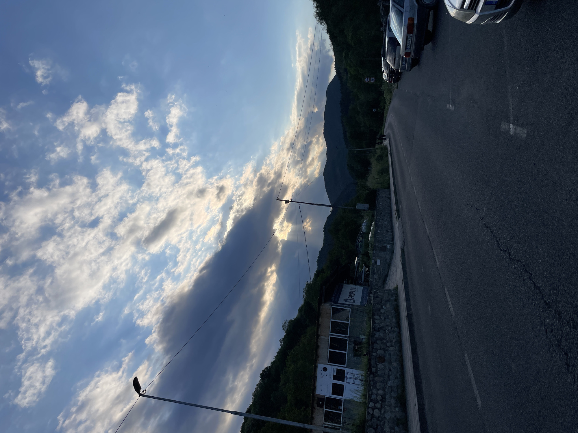The image depicts a view in Zlatograd, Bulgaria. The scene includes a road running along the right side of the frame, with parked cars lining the edge. A building is present on the left side of the road, with a sign visible on its facade. The background features a landscape with trees and a mountain range. The sky is filled with clouds, with sunlight breaking through in certain areas. Power lines are also visible against the sky. The image appears to be taken during the daytime.
