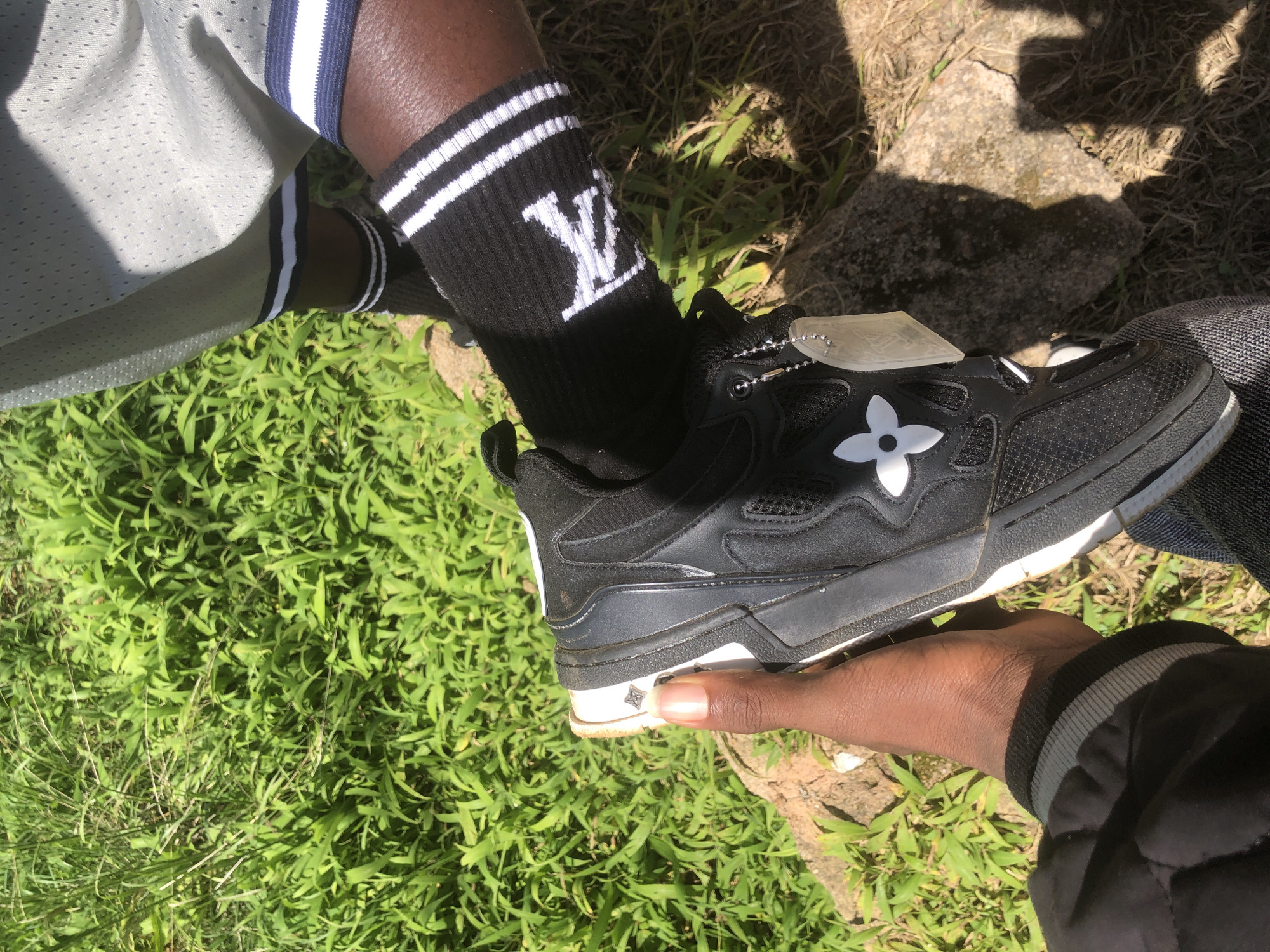 An individual's hand is holding a black and white Louis Vuitton sneaker. The person is wearing black socks with white stripes and the Louis Vuitton logo. They are also wearing grey athletic shorts with white and blue stripes, and a black jacket. The person appears to be in an outdoor, grassy area, possibly in Gwafan, Nigeria. A rock is visible in the foreground.