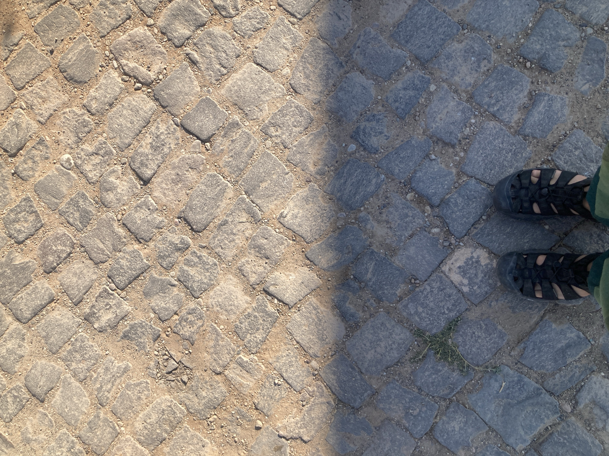 In Avanos İlçe Merkezi, Turkey, a top-down view captures a cobblestone ground textured with sand and small pebbles. The illumination suggests a sunny day, casting a shadow that divides the scene into two distinct lighting zones. In the lower section, two feet are visible, clad in black, open-toed sandals. The left foot is angled slightly outward, while the right foot is positioned straighter. The cobblestones are dark gray in the shadowed area and transition to a lighter, warmer tone in the sunlit upper portion of the image. A small patch of green grass is visible in the lower left quadrant, growing between the stones.