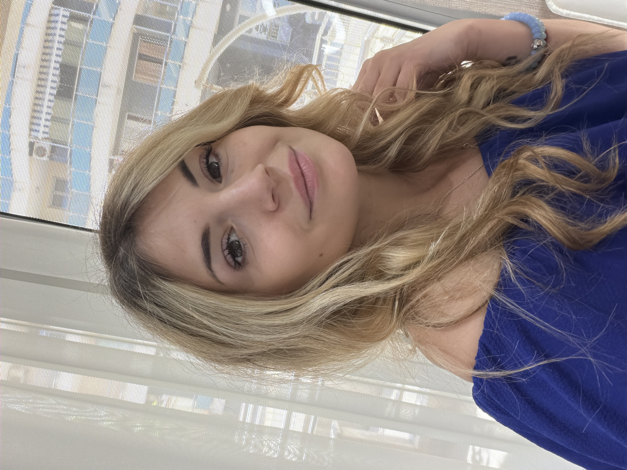 A young woman with blonde wavy hair is seen indoors, wearing a blue shirt and a blue beaded bracelet. She is positioned near a window, through which a modern building with blue and white facade elements is visible. The window has a white mesh blind.