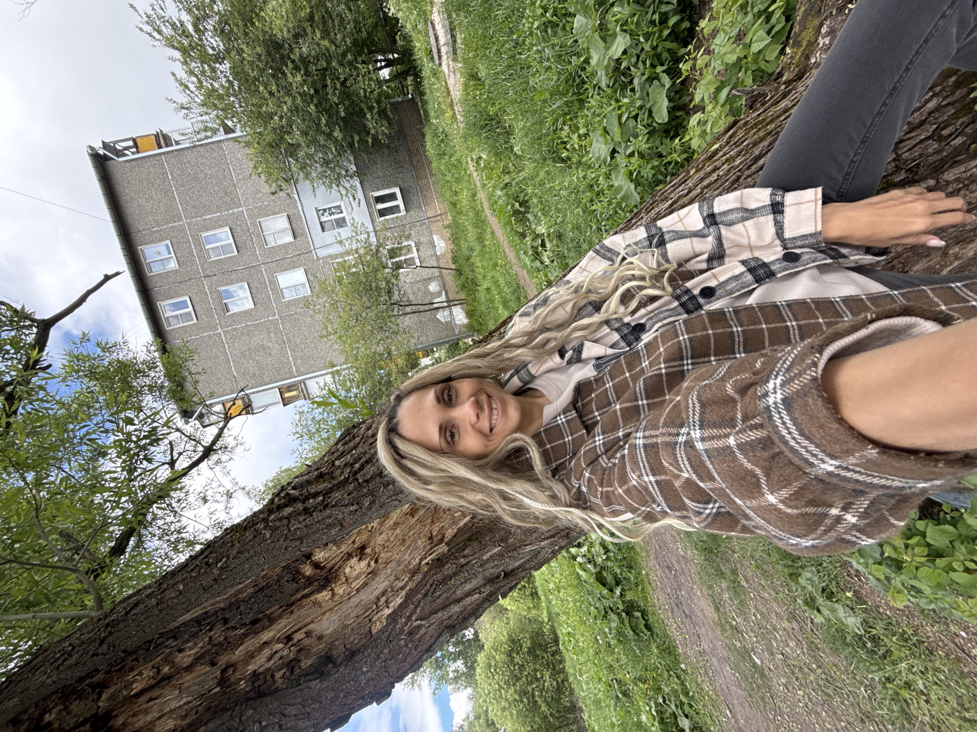 A woman lies on a fallen tree trunk. She is wearing a brown and tan plaid shirt, gray pants, and has blonde hair. In the background is an apartment building, green grass, and trees. The location is likely Заволжье, Russia, based on request.