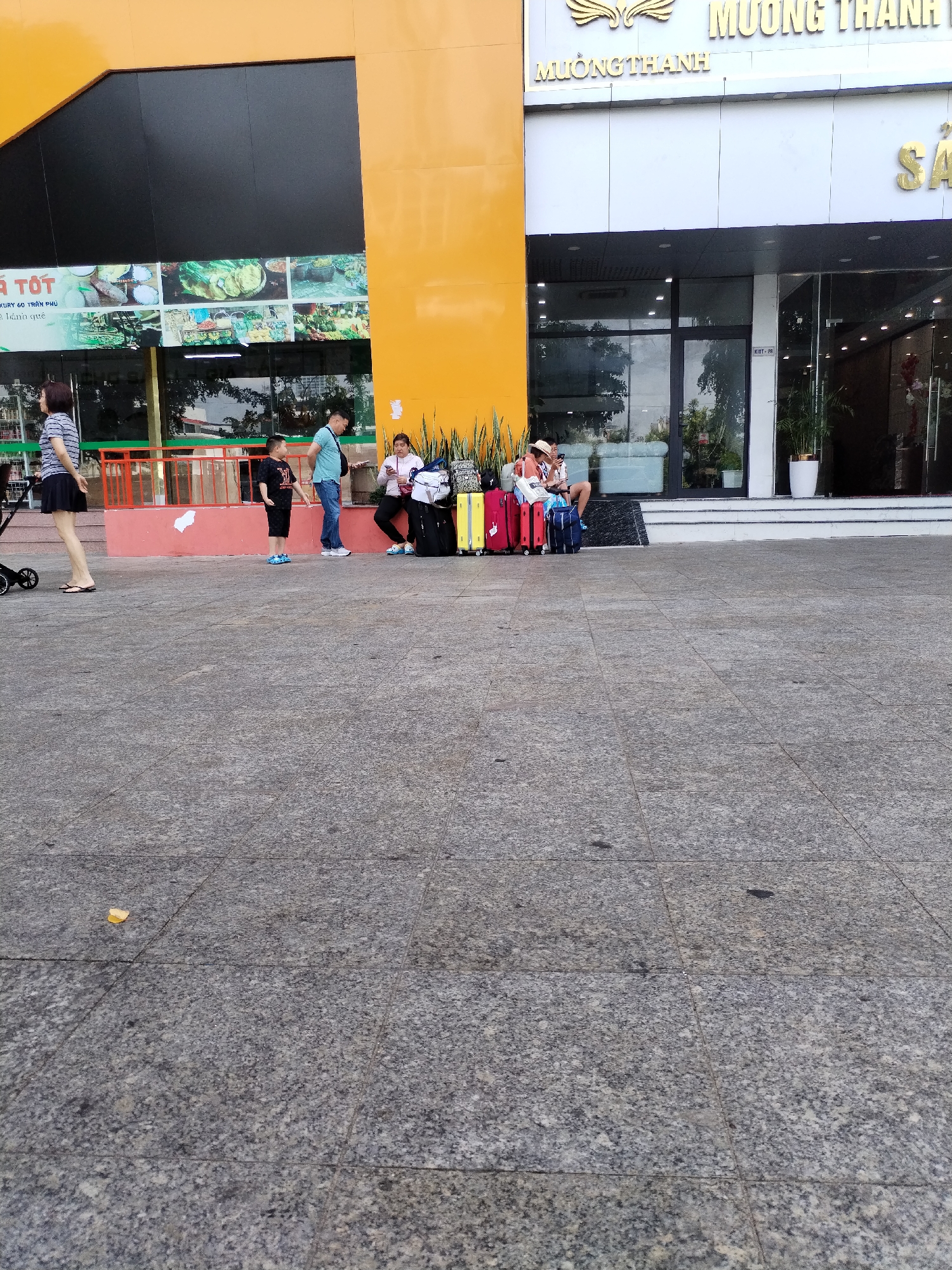 A group of people are gathered on a paved outdoor area in front of a building with a large yellow facade and a glass entrance. On the left, a woman in a striped shirt and black skirt walks a stroller. To her right, a young boy in a black t-shirt and shorts stands. Further to the right, a man in a blue t-shirt and jeans is talking to a woman in a pink jacket who is sitting on a ledge with a young boy wearing a straw hat. They are surrounded by luggage, including red and yellow suitcases and a blue duffel bag. The building appears to be a hotel or commercial establishment, with signage that reads "MUONG THANH" in gold letters. The ground is a grey, speckled paving stone.