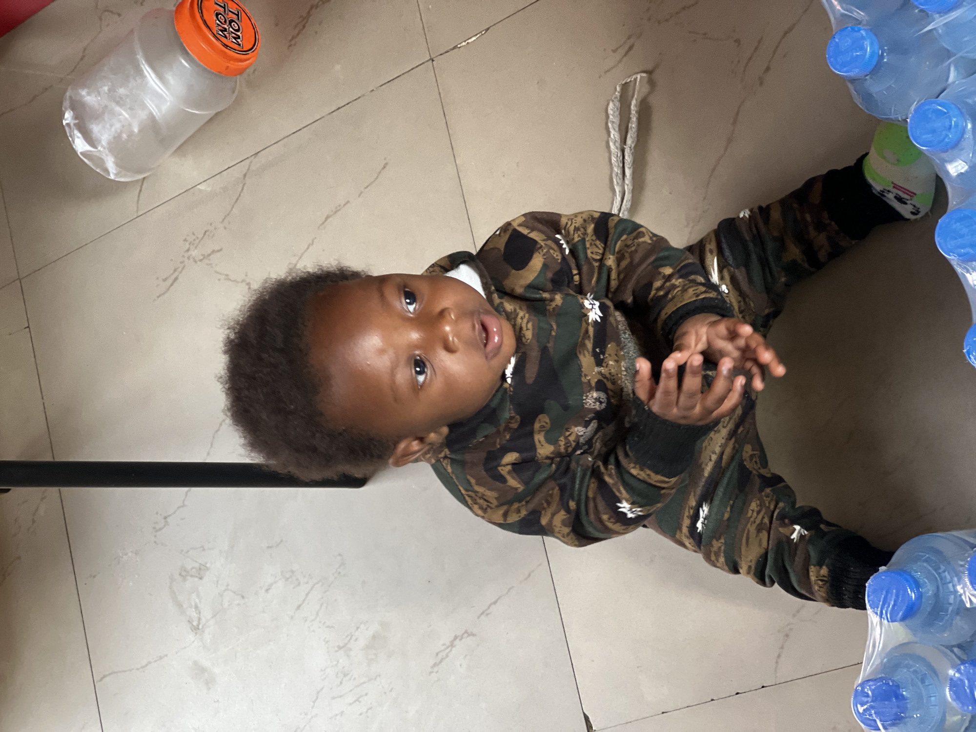 The image, captured in Jos, Nigeria, shows a young child lying or sitting on a light-colored tiled floor, looking directly up at the camera with a curious and engaged expression. The child is wearing a camouflage-patterned long-sleeved top and matching pants. One foot, clad in a light green sock with a white pattern, is visible near a large, shrink-wrapped pack of several plastic water bottles, each with a blue cap.
To the upper left of the child, a clear plastic jar with an orange lid rests on the floor. The text "TOM TOM" is clearly visible on the orange lid. A white braided string or rope can also be seen on the floor, near the child's right side. The overall setting appears to be indoors, likely a home or a storage area, with a smooth, possibly marble or ceramic, tiled floor. The child's hands are slightly open and raised, contributing to an impression of playfulness or interaction with their surroundings. The lighting suggests a daytime indoor scene.