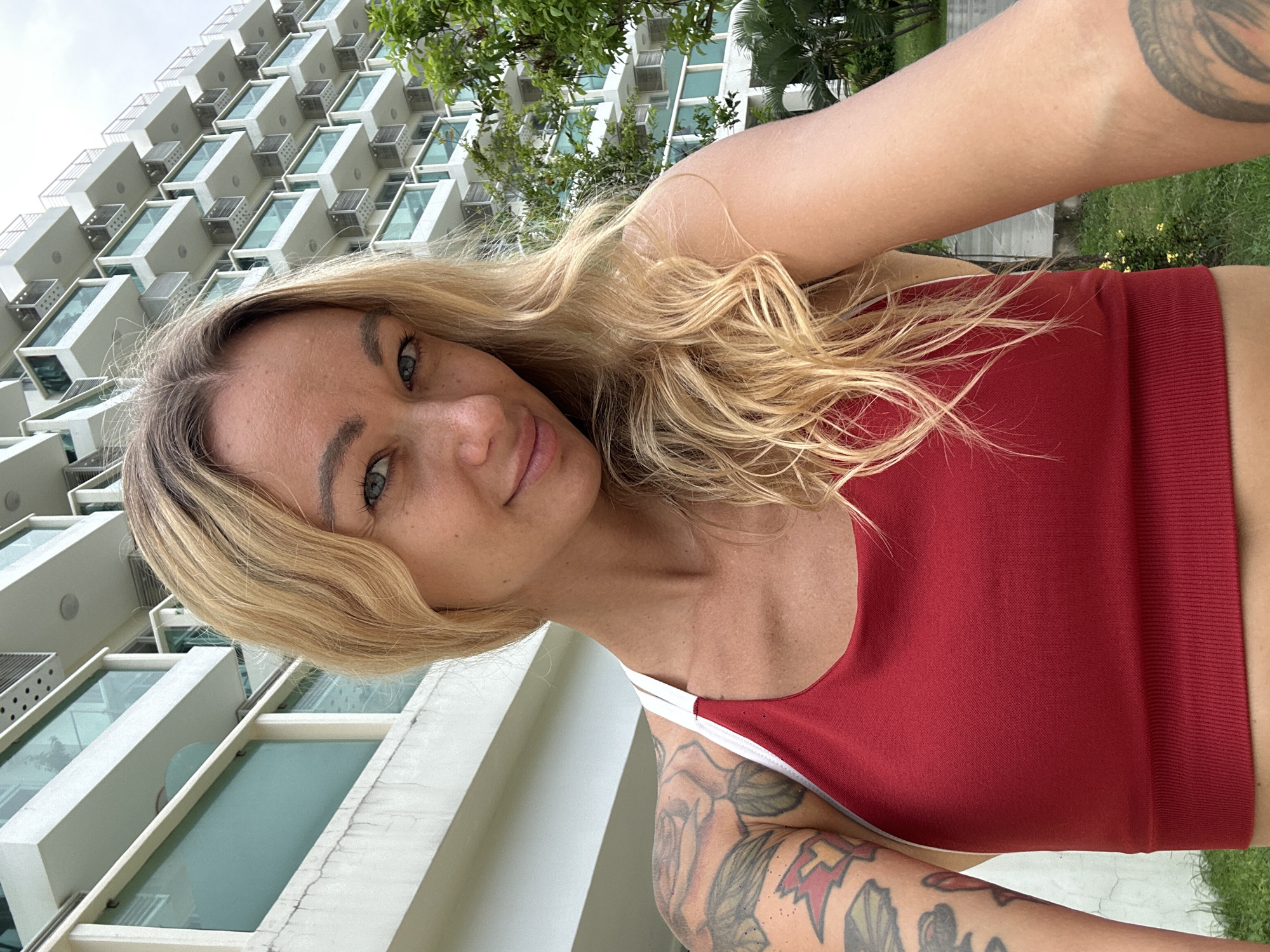 The image shows a woman with blonde hair and blue eyes, wearing a red sports bra. She has tattoos on her left arm. She is standing outside a modern, multi-story building with balconies and glass windows. Lush green trees and foliage are visible in the background. The sky is overcast. The location appears to be a hotel or residential complex.