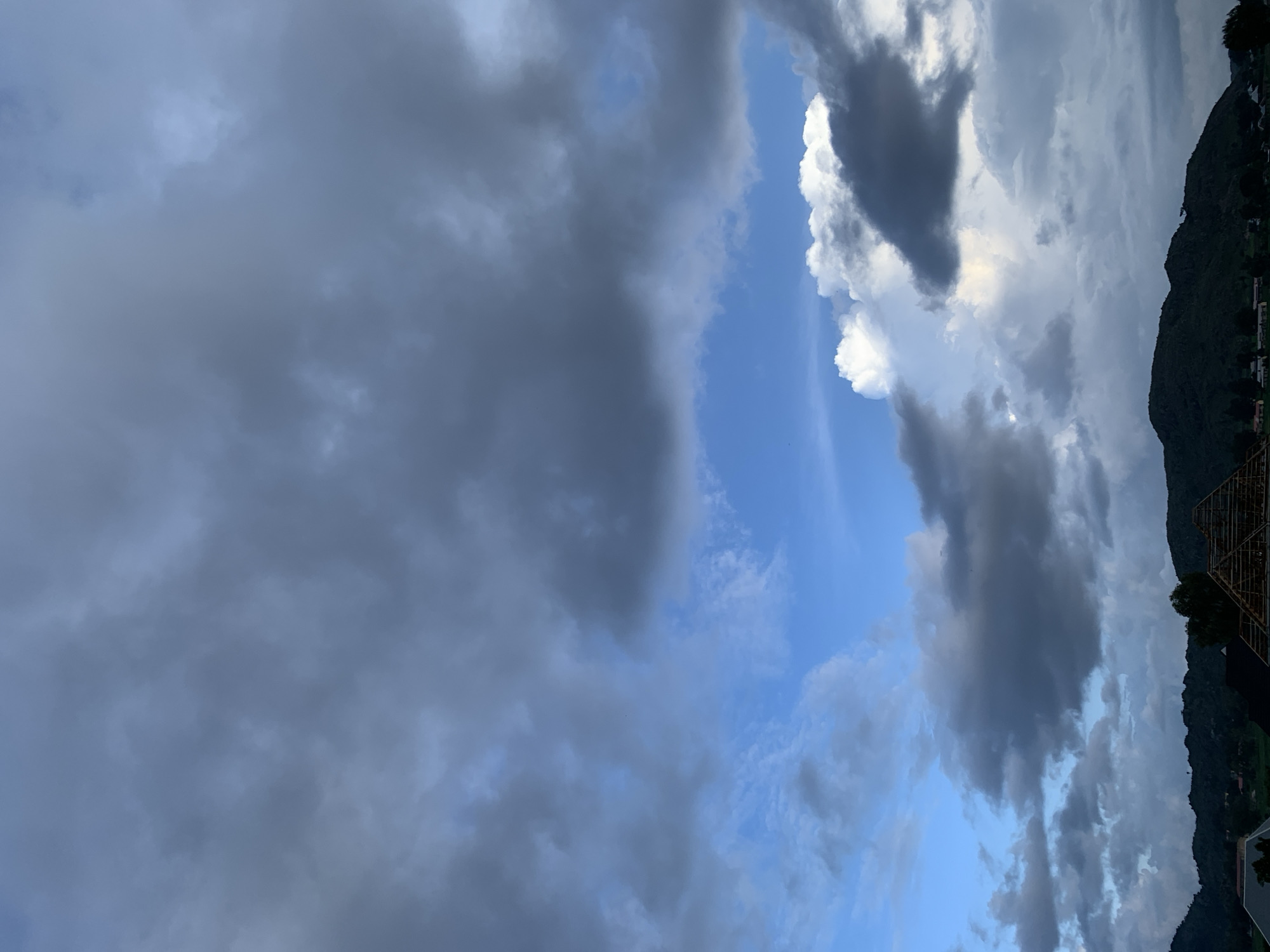 The image depicts a dramatic sky over a rural landscape, likely Kuru Karama, Nigeria, with a prominent hill or mountain range in the background. The sky is filled with a mixture of dark, heavy clouds and patches of bright blue sky, suggesting an approaching storm or a clearing after rain. The clouds are predominantly dark gray and appear dense, with some lighter, fluffier clouds interspersed.
In the foreground, the silhouette of a landscape is visible against the sky. This includes a dark, undulating hill or mountain line, with hints of vegetation. Below the hill, there are some scattered structures, possibly houses or buildings, with some appearing to be under construction, evidenced by exposed roofing frameworks. There are no people or animals clearly visible in the image. The overall impression is one of a natural, somewhat moody atmospheric scene in a rural setting.