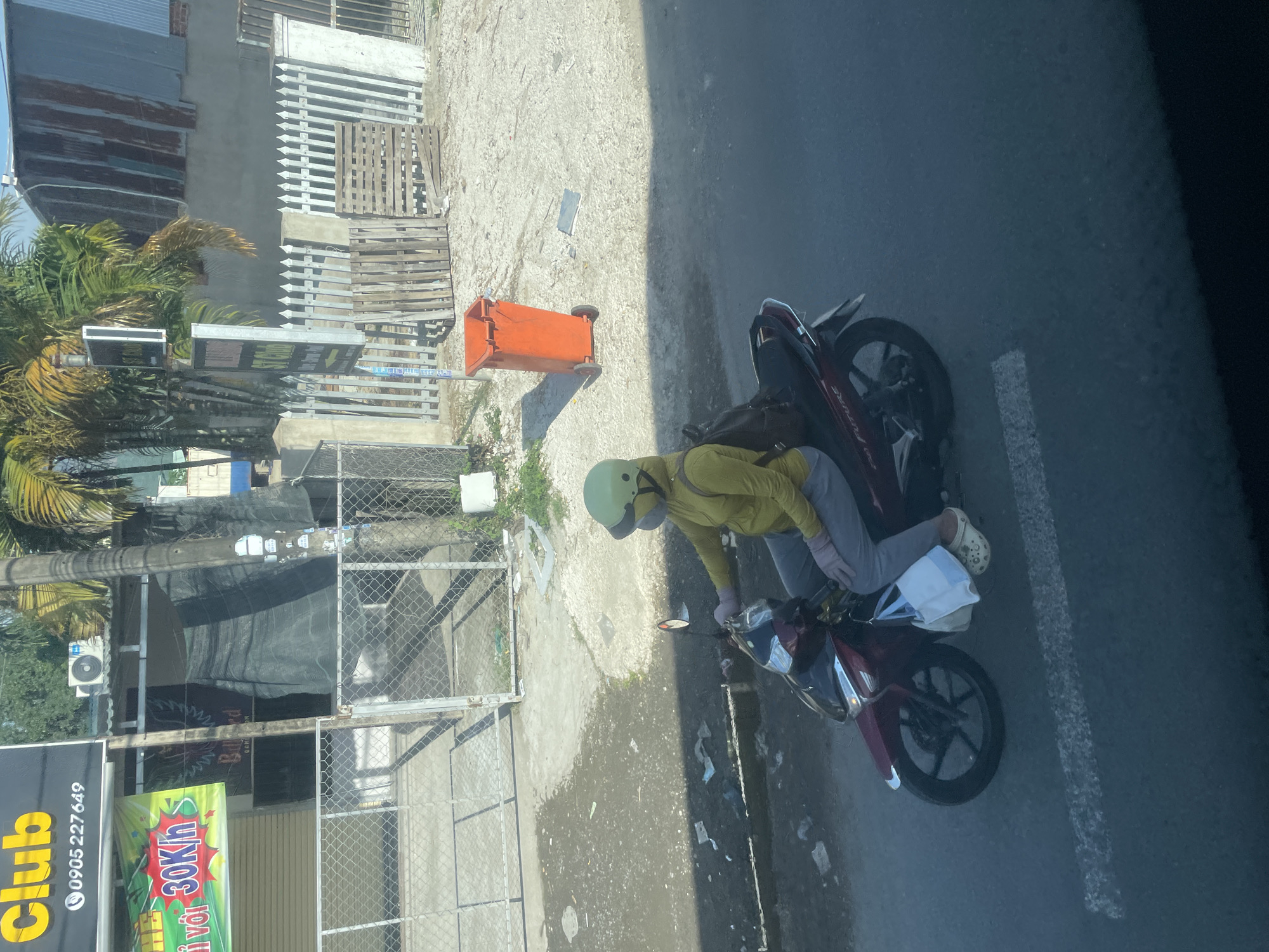 The image shows a person riding a red motorcycle in what appears to be a city street in Tay Nha Trang Ward, Vietnam. The rider is wearing a yellow long-sleeved shirt, grey pants, a light green helmet, and pink gloves. They are also wearing a mask that covers their nose and mouth, and their face is obscured. A black backpack is strapped to their back, and a white bag with blue trim is hanging from the handlebars. The motorcycle is a scooter-type with a front disc brake. The rider's feet are shod in white clog-style shoes.
The background reveals a street with a paved surface and some scattered debris, including paper and plastic. To the left, there is a building with a black sign that has yellow lettering. Below this sign, there is another sign with a colorful graphic and the text "30Kh". A metal fence runs along the edge of the property. Further back, there are palm trees and a building with a corrugated metal roof. To the right, an orange trash bin is visible next to a white picket fence and some wooden pallets. The overall scene suggests an urban environment, possibly during daylight hours, with signs of everyday activity and some litter on the ground.