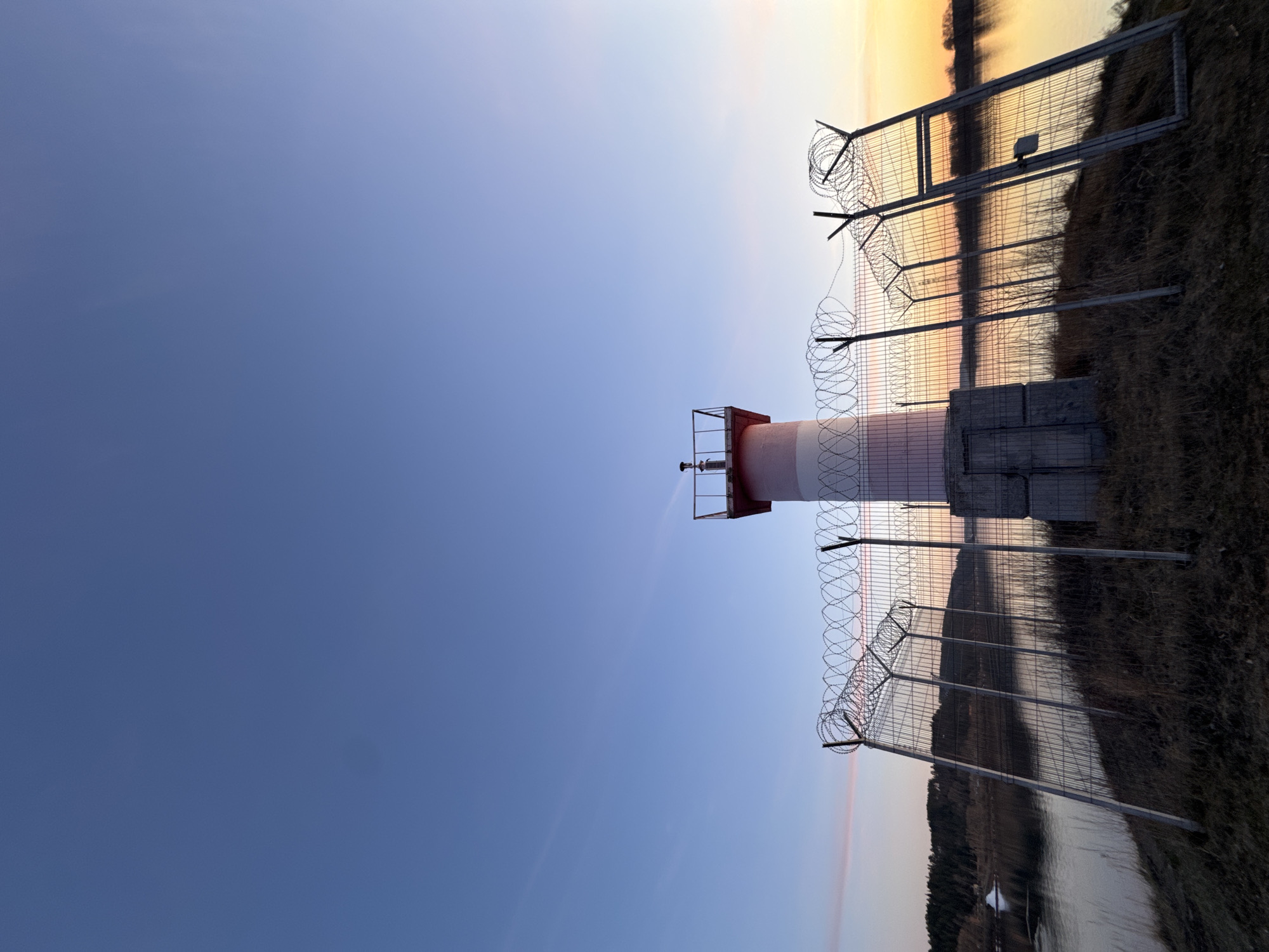The original image, presented in a vertical orientation, captures a serene landscape during what appears to be either sunrise or sunset. The scene features a clear sky, a body of water, and a prominent red and white striped structure that functions as a lighthouse or navigational beacon.
The beacon, painted in alternating red and white bands, is anchored on a base made of concrete blocks. Near its top, a small metal platform is visible, likely holding the light or sensor.
A sturdy metal fence, reinforced with multiple coils of razor wire along its top, traverses the midground. This barrier runs parallel to the body of water, suggesting a secured or restricted area. Beyond the fence, the calm surface of the water reflects the warm hues of the sky.
The sky exhibits a clear gradient, transitioning from a deep blue at the top and left to a brighter, yellowish-orange glow near the horizon on the right. This warm light illuminates the scene, casting reflections across the water. On the far side of the water, dark landmasses or distant foliage are faintly visible.
There are no people or discernible activities taking place in the image, contributing to a tranquil and possibly isolated atmosphere. No text is visible within the frame. The setting suggests a riverside or reservoir location, consistent with the given context of Chaikovsky, Russia.