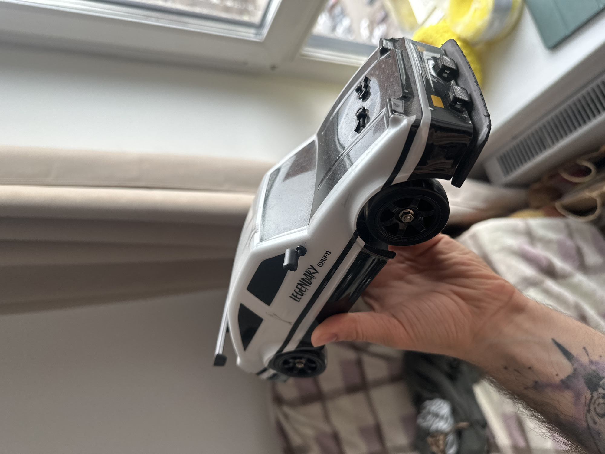 The image shows a person holding a toy car indoors. The car is white with black accents and the words "LEGENDARY (DRIFT)" printed on its side. The person holding the car has tattoos on their arm. The background includes a window, radiator, and furniture. The location in the city of Lyubertsy, Russia, is not evident from the image.