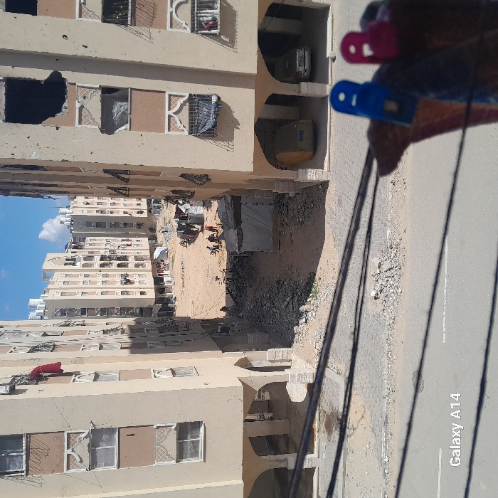 The image, marked "Galaxy A14" in the lower-left corner, depicts an urban residential scene in the city of Khan Yunis, Palestinian Territory, under a bright blue sky with scattered white clouds. In the immediate foreground, a paved road is visible, partially obscured by three horizontal dark cables. To the far right, a hand-held object with a blue and a pink clothespin grips a piece of reddish-brown fabric.
The midground features two multi-story, light-tan residential buildings that frame a central open space. The building on the left exhibits minor facade wear, with a red garment hanging from an upper window. The building on the right is extensively damaged, featuring several large, irregular holes exposing interiors in its upper floors and numerous broken windows with visible internal debris. Two vehicles, one covered in yellow and another white, are parked within its ground-floor arched structures.
The central open area between the buildings consists of unpaved sand and dirt, with scattered piles of rubble. A group of approximately five to seven individuals, dressed in casual clothing, are observed in this central area, appearing to engage in excavation or sorting activities amidst debris and a small, white tarp-covered structure. In the background, additional multi-story residential buildings of similar construction are visible, some also displaying signs of damage and topped with numerous white water tanks.