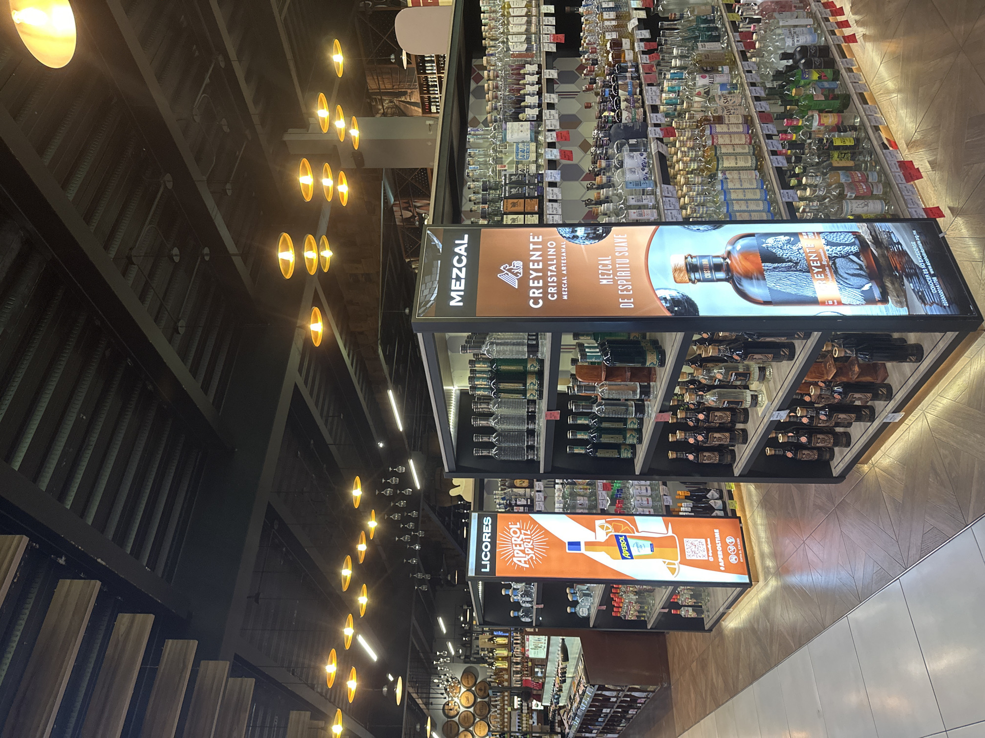 The image captures an interior view of a store, seemingly a liquor store, with shelves stocked with various alcoholic beverages. Banners advertising specific brands, such as "Mezcal Creyente" and "Aperol Spritz," are prominently displayed. The environment features a high ceiling with rows of suspended lights, contributing to the store's illumination. The floor has both wood and white tiled sections. Based on the OCR text, the image suggests a location in Puebla, Mexico.