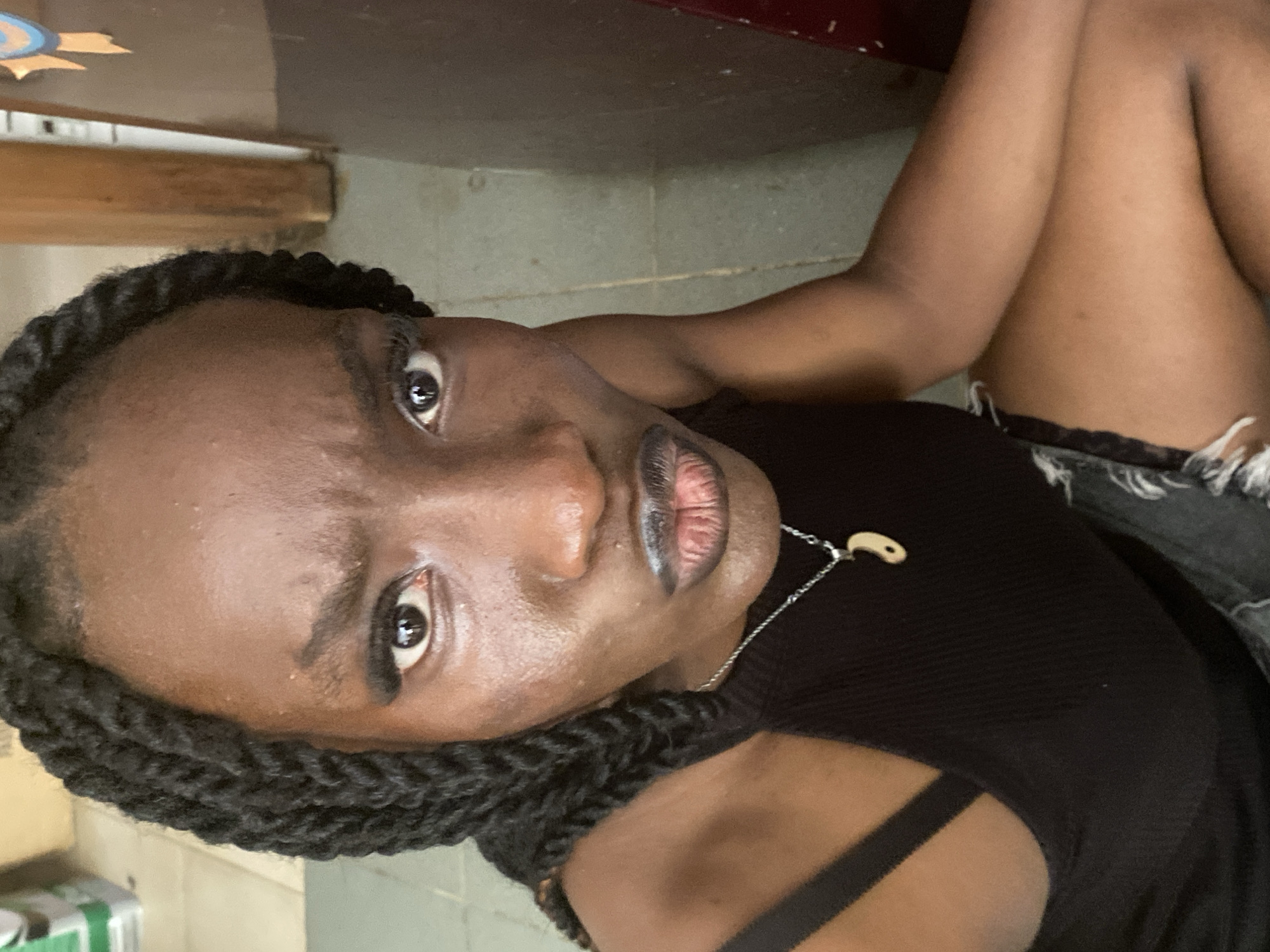 A close-up, high-angle, medium shot shows a young Black woman with braided hair, dark lipstick, and a black tank top. She is looking directly at the camera with a pouty expression. Her left arm is bent, and her right arm is resting on her leg. The background is out of focus and appears to be indoors, with a tiled floor and a wooden cabinet. The scene is likely within a home or shop in Iju, Nigeria.