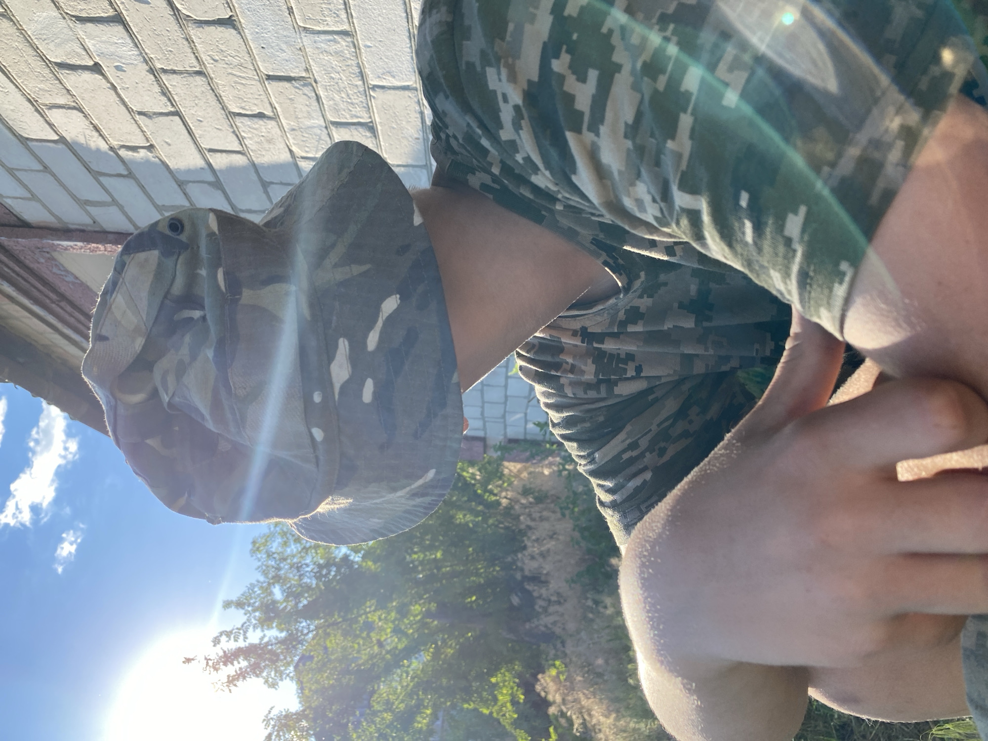 A person wearing a camouflage hat and shirt sits outdoors next to a brick building with a red-trimmed roof. The sun is bright in a blue sky with clouds, and trees and dry grass are visible in the background. The person's face is obscured by the hat. The scene appears to be in a rural or suburban setting.
