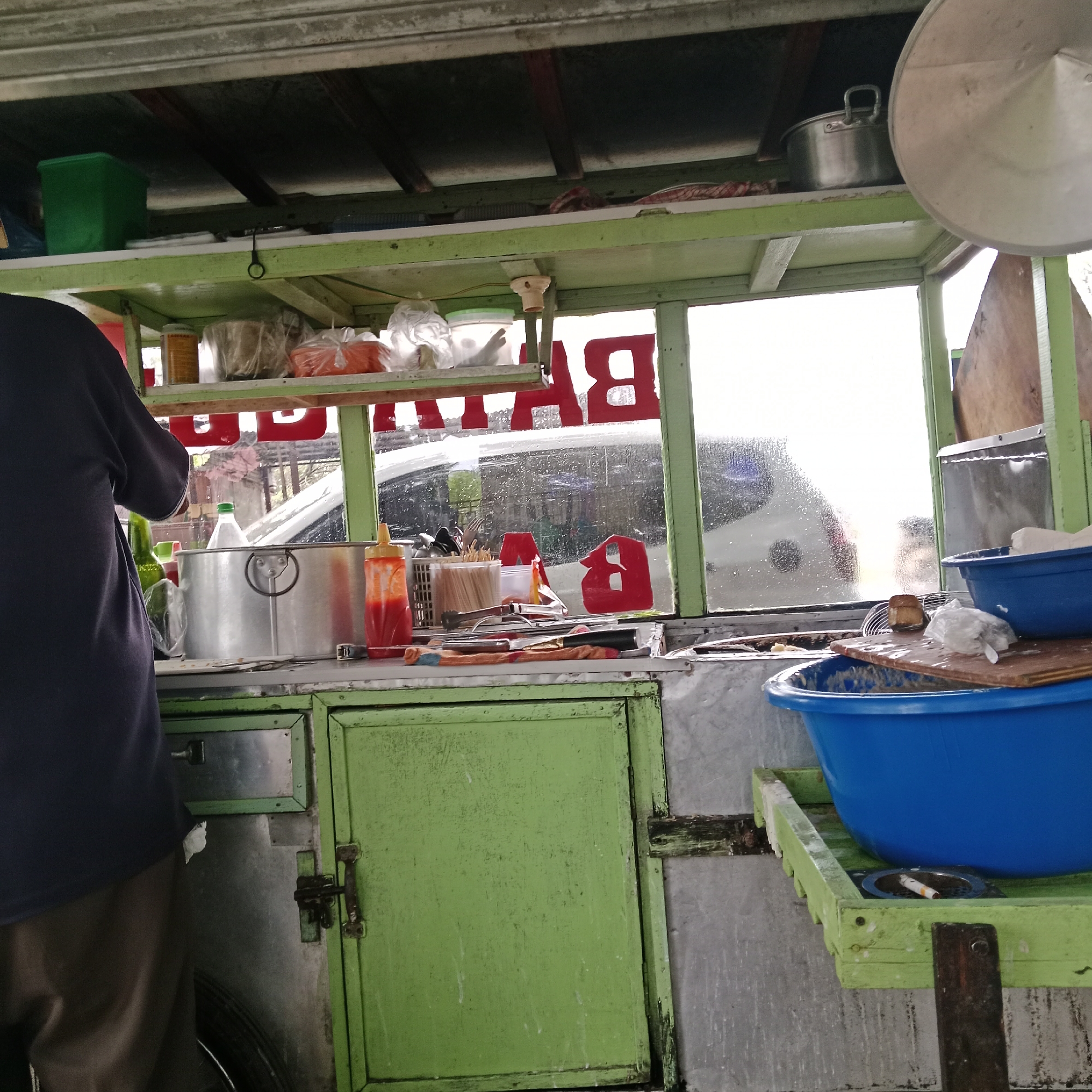 An outdoor food cart is depicted from a low angle, showcasing a person in a dark blue shirt standing on the left, facing away from the camera. The cart itself is a mix of green-painted wood and metal. A shelf above the counter holds various items, including a green plastic container, bags of food, and a metal pot. The counter area features a large metal pot, a bottle with a green cap, a ketchup dispenser, a crate of wooden skewers, and other kitchen utensils. Red lettering, possibly part of a sign, is visible on the glass window behind the counter. Outside the window, a blurry white vehicle is visible. Below the counter, green painted cabinets are present, with one door ajar. On the right side of the cart, a wooden platform holds a large blue plastic basin. The scene appears to be outdoors, possibly on a street, with a weathered and utilitarian aesthetic.