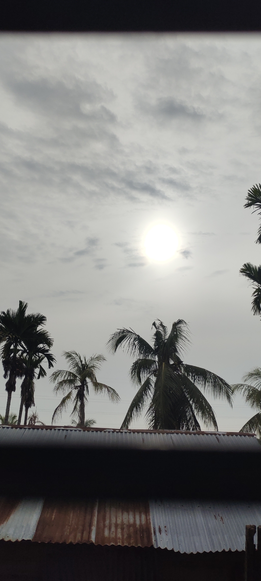 The image captures a bright, overcast sky with the sun appearing as a luminous disc partially obscured by clouds. Several palm trees are silhouetted against the sky, their fronds creating a textured canopy. In the foreground, a corrugated metal roof, exhibiting signs of rust and weathering, occupies the lower portion of the frame. The scene appears to be from Lhokseumawe, Indonesia, suggesting a tropical or subtropical climate. There are no people or discernable actions depicted. The overall mood is one of a cloudy day with diffused sunlight.