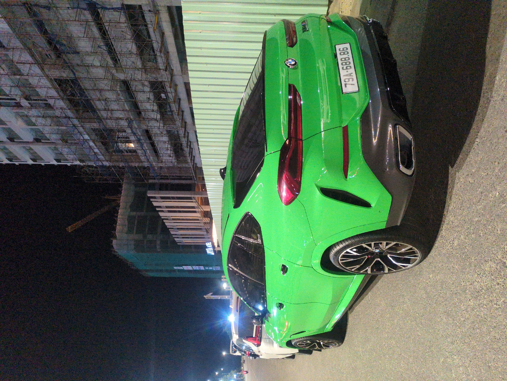 The image shows a bright green BMW X6 with license plate 79A-688.86 parked on a street in South Quy Nhon Ward, Vietnam at night. In the background are tall buildings, one under construction with visible scaffolding and a crane. Other cars are parked further down the street. The overall lighting is dark, illuminated by street lights and car headlights.