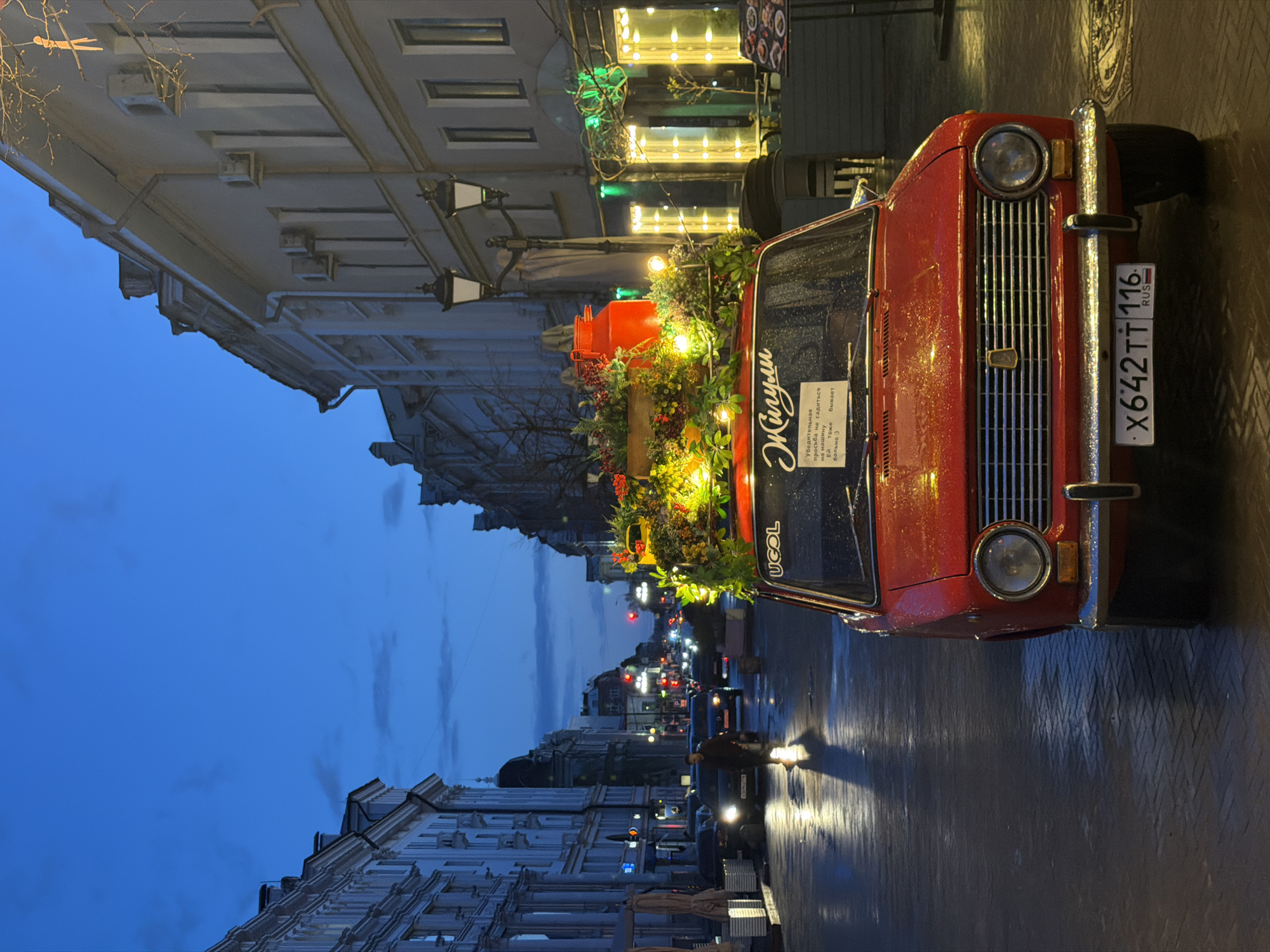 The image depicts a street scene at dusk or early night in Kazan, Russia, after recent rain, as indicated by the wet pavement reflecting streetlights. The primary subject is a vintage red Lada (Жигули in Russian) car, parked on the side of the road.
The car is elaborately decorated with garlands of greenery, berries, and numerous small, warm-toned lights, creating a festive appearance. A large red bucket or planter is also visible on the car's hood, nestled within the decorations. On the car's windshield, there's a sign with text in Russian that reads: "Жигули" (Zhiguli, the car brand), and below it, "Убедительная просьба не садиться на машину. Ей тоже бывает больно :)" which translates to "Urgent request: do not sit on the car. It also gets hurt :)". Another visible text on the upper part of the windshield, though partially obscured and upside down in the original orientation, appears to be "UGOL". The car's Russian license plate, X642TT 116 RUS, is clearly visible on the front bumper. The "116" region code corresponds to the Republic of Tatarstan, where Kazan is located.
The street is lined with multi-story buildings featuring classical architectural elements. On the right side, bright lights from what appears to be a cafe or shop illuminate the sidewalk. Several old-fashioned streetlights with multiple lanterns are also present. In the background, other cars are visible, their headlights and taillights creating streaks of light. A person can be seen walking on the wet pavement, illuminated by the lights. The sky is a deep twilight blue, transitioning to darker hues overhead. The overall atmosphere suggests a lively urban setting, perhaps an evening out or a special display on the street.