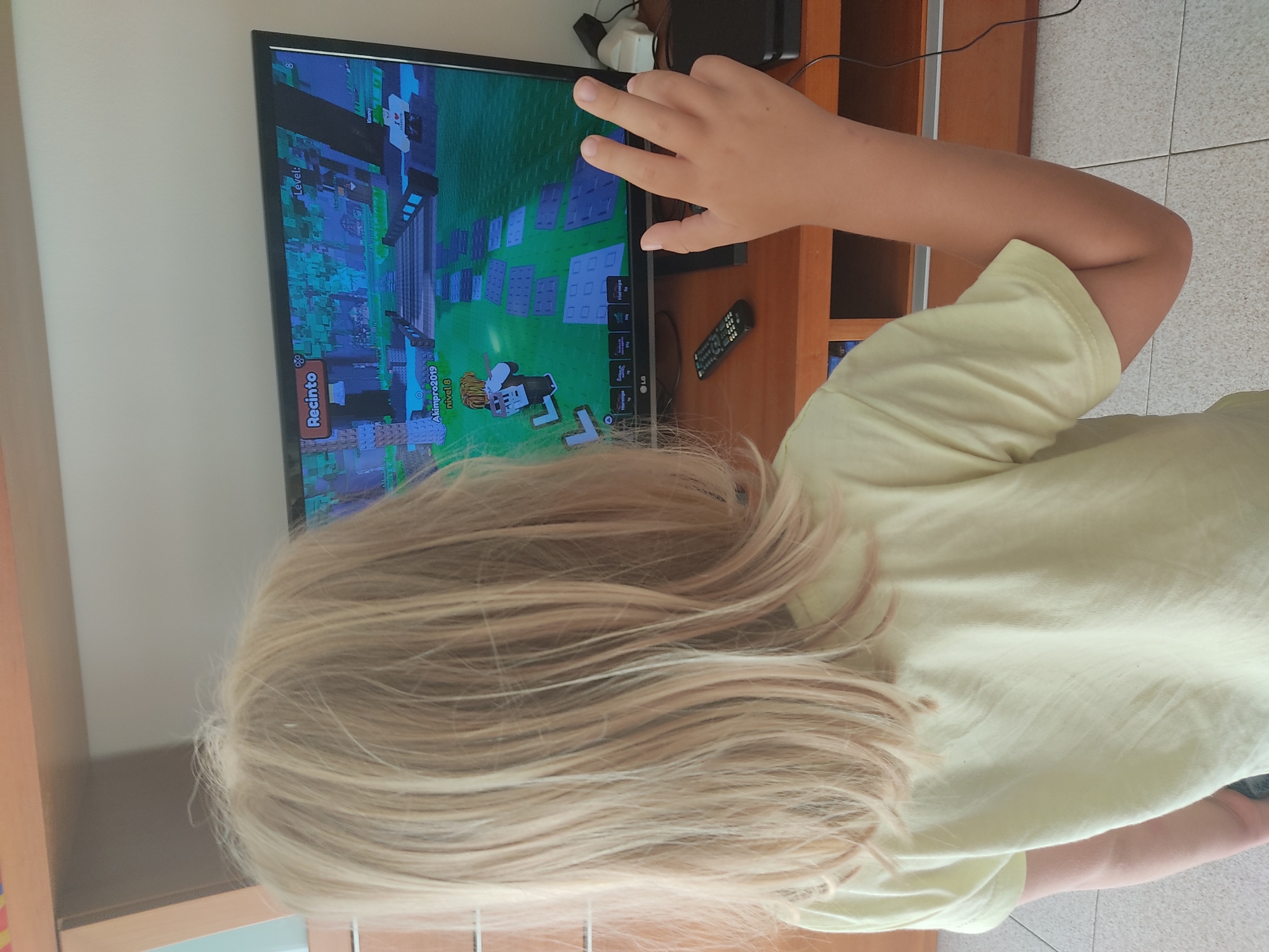 A young person with long blonde hair is standing with their back to the camera, facing a computer monitor that displays a video game. The game appears to be a blocky, 3D environment with a pathway leading into a forest. Text on the screen indicates "Recinto" and "Level: 8". The person is wearing a light yellow t-shirt and has their right hand raised, palm facing forward, with four fingers visible. On the wooden desk below the monitor, a black remote control and some electronic devices are present. The scene takes place in an indoor setting, likely a home, with shelving units visible in the background. The floor is tiled with a light grey and white pattern. The specific location of Blanes, Spain is not discernible from the visual information provided in the image.