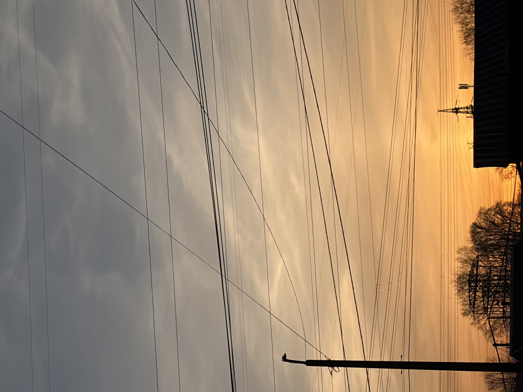 The image depicts a sky at sunset in what appears to be an urban or industrial area. The sky transitions from a grey color on the left to a golden-orange hue on the right, suggesting a sunset. Numerous power lines crisscross the frame, creating a dense network against the sky. On the right side, there are silhouettes of trees and what appears to be a metal structure, potentially a power transmission tower or building framework. The presence of power lines and structures suggests an industrial or suburban area. Without specific landmarks, pinpointing the exact location within Chaikovsky, Russia is not possible. The scene appears to be still, capturing a moment of twilight.