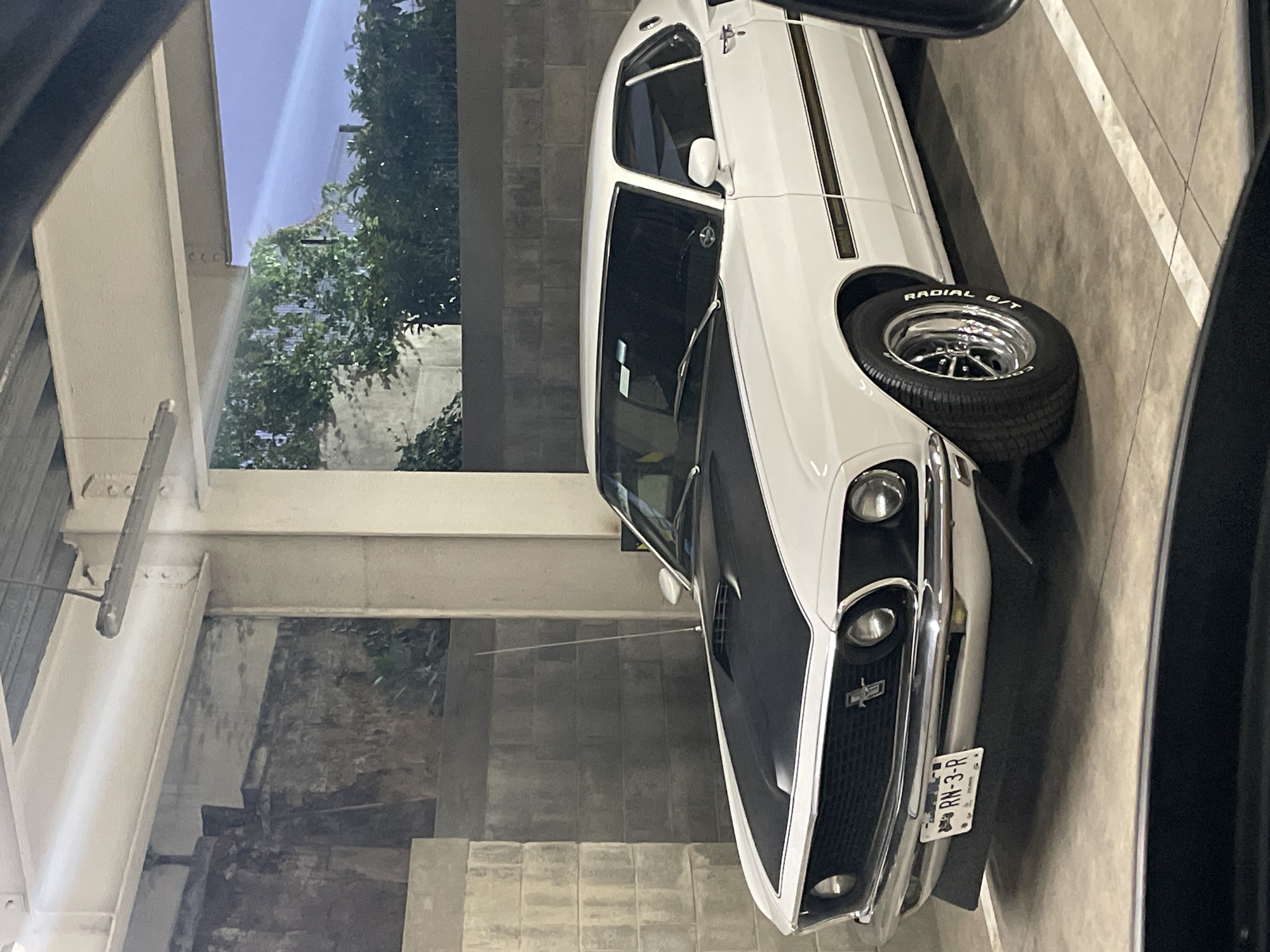 A white 1969 or 1970 Ford Mustang Mach 1 is parked in an indoor parking garage in Huixquilucan de Degollado, Mexico. The car has a black hood and racing stripes, and its license plate reads "RN-3-R". The environment is a concrete parking garage with trees visible in the background. The photo appears to have been taken from inside another vehicle.