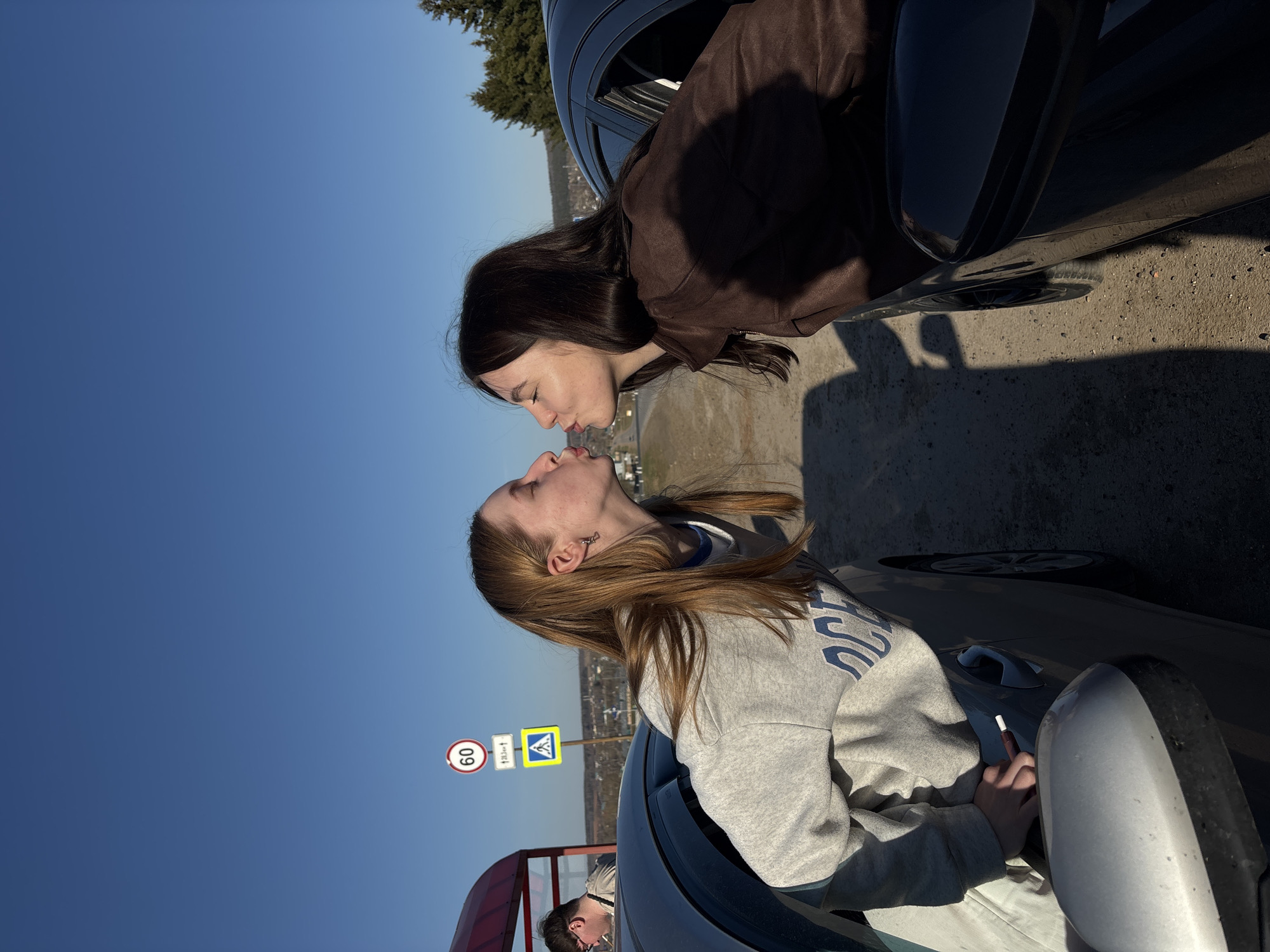 Two women are in a parked car. One woman is looking out of the driver’s side window; she has light-brown hair and is wearing a light-grey sweatshirt. The other woman is in another car next to her; she has dark hair and is wearing a brown jacket. The women are very close to each other, their lips touching. There is a 60km speed limit sign and a crosswalk sign in the background. The location appears to be Баклуши, Russia.
