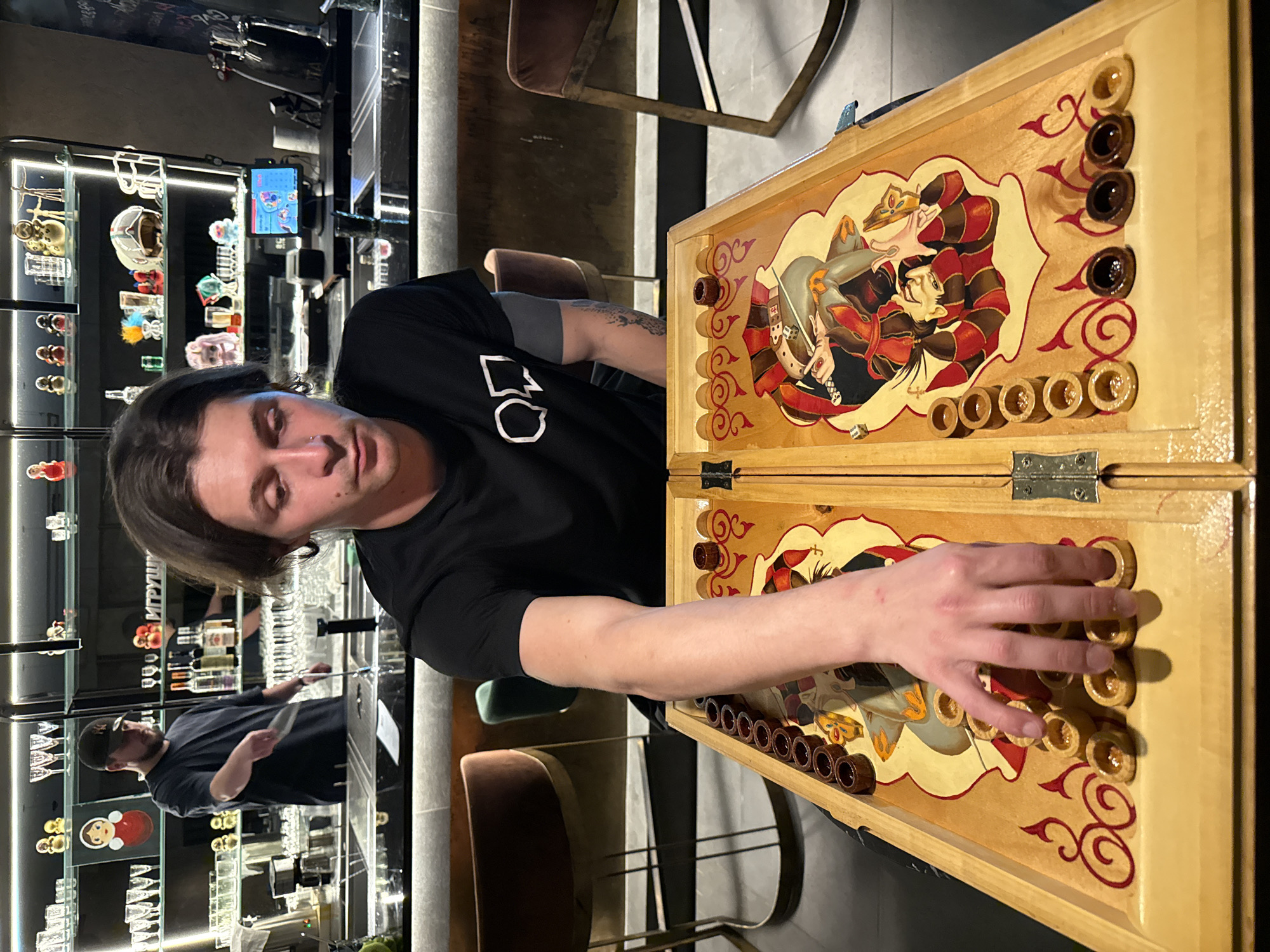 A man is playing a game of backgammon in a dimly lit indoor location, possibly a bar or restaurant in Sochi, Russia. The backgammon board has a painted design featuring figures. The man is wearing a black t-shirt and has short dark hair. Another man, partially visible, is standing behind a bar with various items displayed on shelves.