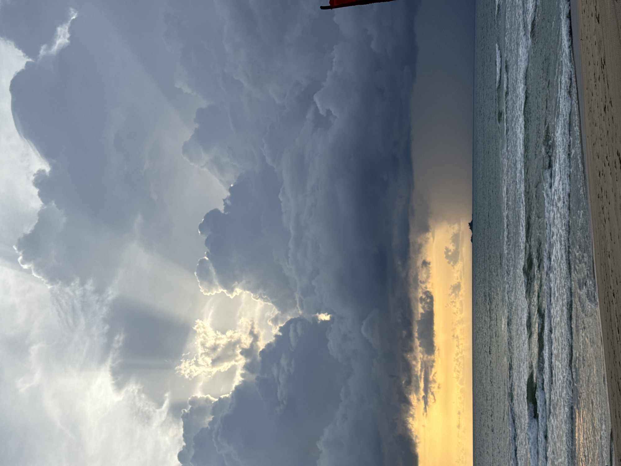 The image captures a dramatic sky over a beach scene. The sky is filled with dark, heavy storm clouds, but a bright sliver of sunlight breaks through, creating visible sunbeams that fan out. This contrast between the darkness and light suggests an approaching storm or the clearing after one.
To the right of the sky, the ocean is visible, with gentle waves lapping at the shore. The water appears a muted grey-blue, reflecting the overcast sky. The foreground features a wooden structure, possibly a boardwalk or pier, running vertically along the right edge of the frame. The texture of the wood is rough and weathered, hinting at its outdoor exposure.
The overall mood is somewhat somber yet awe-inspiring, due to the powerful display of natural elements. The time of day is likely late afternoon or early evening, given the angle of the light and the colors in the sky. While the location is identified as Choeng Thale, Thailand, there are no specific landmarks or text visible within the image to further pinpoint the exact spot. The image does not contain any people or explicit objects beyond the natural scenery and the wooden structure.