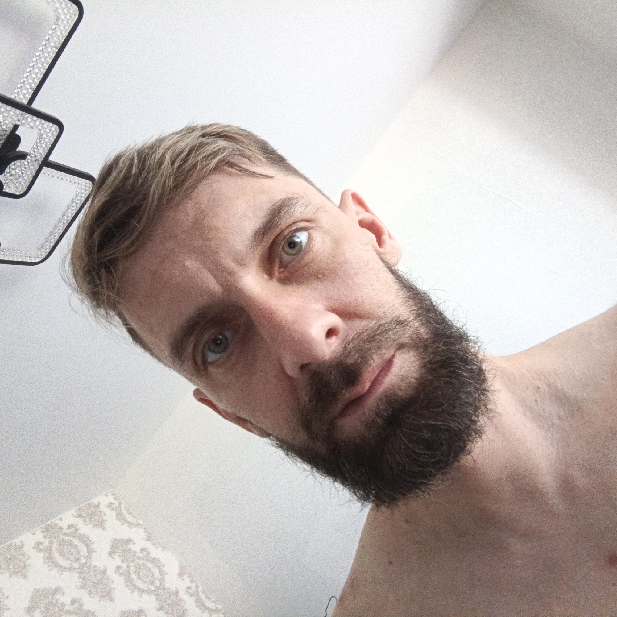 A man with a beard and light brown hair is shown in a close-up selfie. The camera angle is from above, looking down at the subject. The man has light eyes and is looking directly into the camera. The background is a white wall with a patterned section at the bottom left. A modern chandelier with crystal accents is partially visible in the upper left corner. The man appears shirtless. The location is inside a building, possibly a home, in Марковский, Russia.