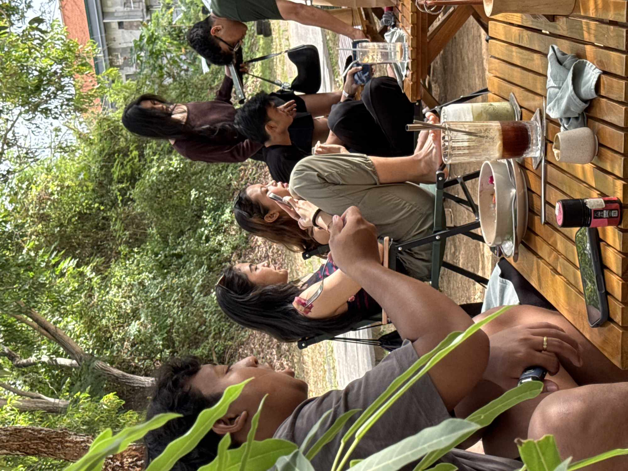 In Kuta Selatan, Indonesia, a group of people are gathered at an outdoor cafe. The foreground features a person in a grey shirt, their arm and hand resting on a wooden table. A ring is visible on their left hand. Next to them on the table are a smartphone, a small beige cup, a folded light blue cloth, and a small black bottle with pink and white labeling.
In the mid-ground, several individuals are seated or standing. A woman in a dark top is looking at her phone. Another woman, wearing a floral top and green pants, is also looking at her phone, holding a fork with food. A man in a black t-shirt is seated, leaning forward and looking at a phone or tablet. Another man in a dark green shirt is standing and looking down at something on a table.
The background consists of lush green trees and foliage, with a building visible behind the trees, suggesting an urban or semi-urban environment. The tables and chairs are simple and functional, typical of an outdoor dining setting. Sunlight filters through the leaves, casting dappled shadows on the ground. The atmosphere appears relaxed, with people engaged in their own activities and interactions.