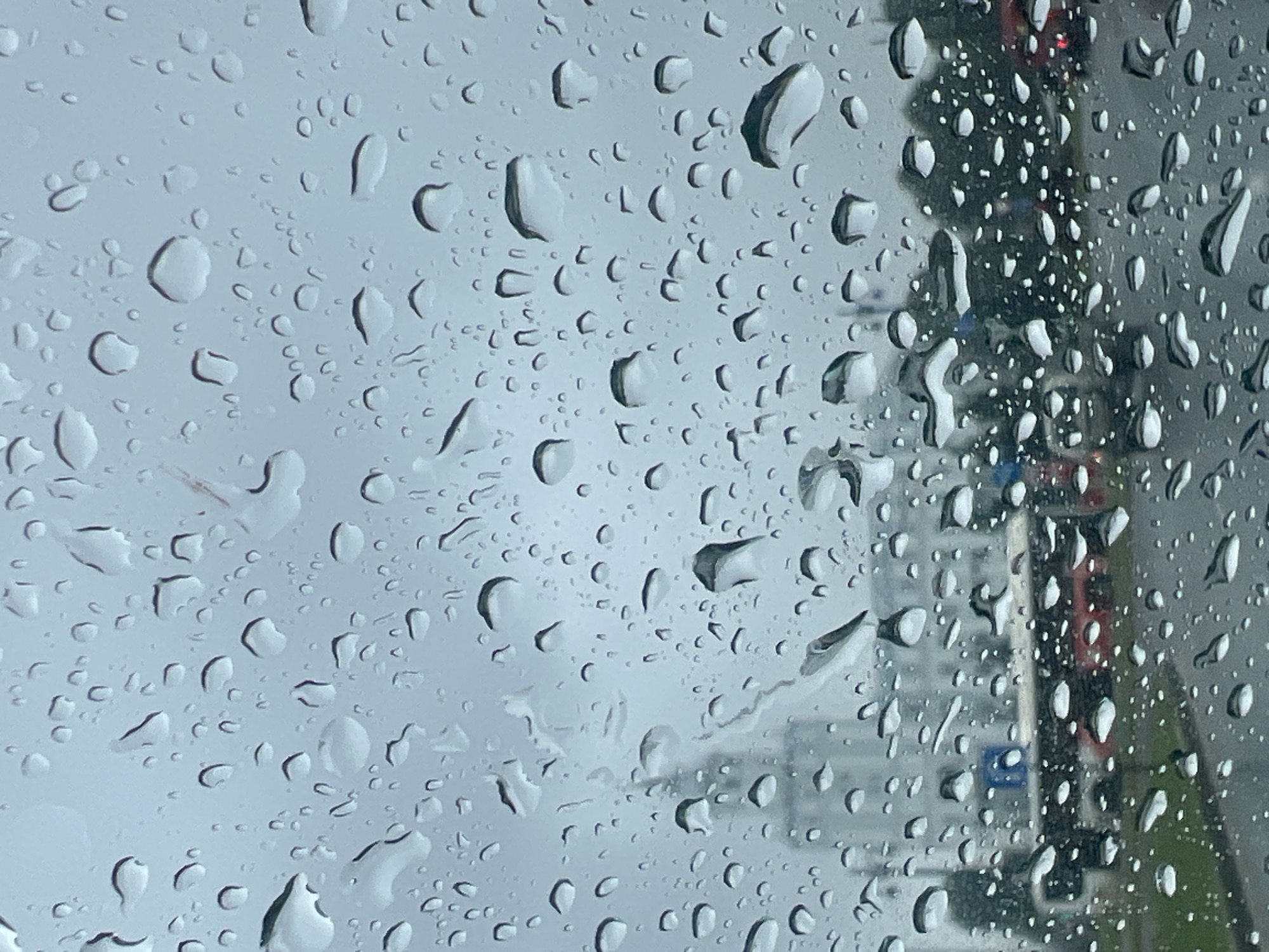 The image, captured on a rainy day, shows raindrops on a glass surface, distorting the view of an urban landscape. In the background, a tall, light-colored building, possibly a government building or a modern office complex, stands against a grey, overcast sky. The architecture suggests a contemporary urban design. Below the building, a red bus is visible, suggesting public transportation is operating. Several cars are also present, indicating a street or road. The environment appears to be a city street with wet asphalt. The scene is consistent with the city of Perm, Russia, given its characteristic architecture and urban infrastructure. There are no visible individuals in the frame. The overall impression is one of a typical rainy day in a Russian city.