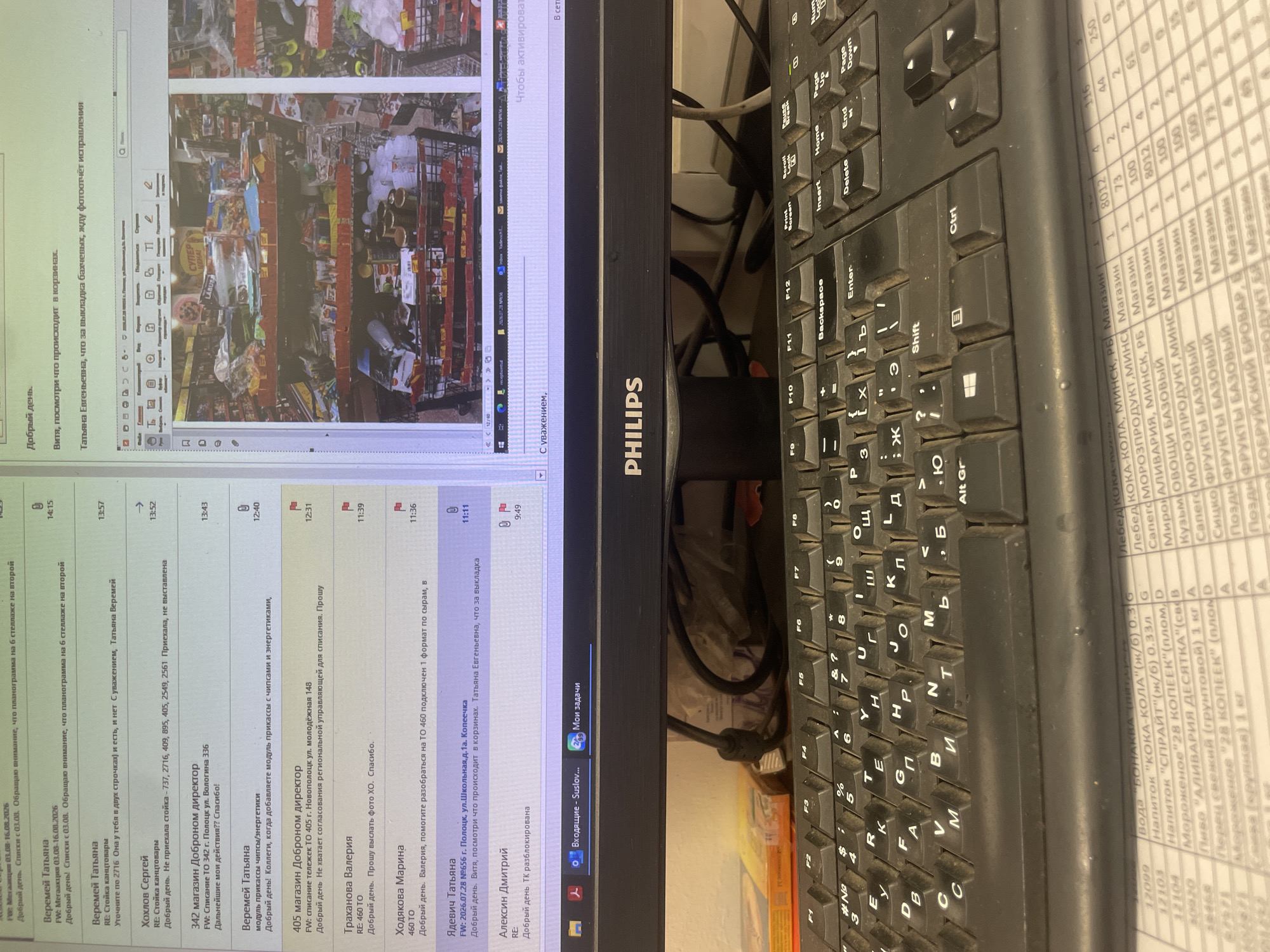 The image shows a computer monitor displaying an email client with a list of messages and a web browser open to an online store, likely within a retail setting. The email client lists several messages with sender names like "Veremey Tatyana," "Hohlov Sergey," and "Trakhanova Valeria," and the content suggests work-related communication regarding merchandise, store operations, and inventory. One email specifically mentions "405 magazin Dobronom director" and a request for a regional coordinating manager's approval for a write-off. Another email from "Yadyevich Tatyana" details a location in "p. Polotsk, ul. Shkolnaya, d.1a, Konechka," indicating a possible address within the Voropaevskiy selskiy Sovet, Belarus.
The web browser displays a visual of store shelves stocked with various goods, including canned items, packaged foods, and beverages. A shopping cart is partially visible, filled with merchandise. The overall environment appears to be an office or backroom area within a retail store, with a keyboard and monitor in the foreground, suggesting the user is actively working. The scene is lit by artificial light.