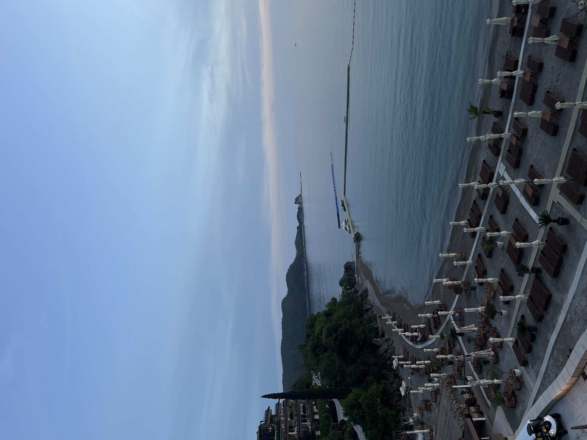 The image captures a high-angle, coastal scene in Budva, Montenegro. The view includes the Adriatic Sea, a beach with rows of loungers and umbrellas, and adjacent land formations with vegetation and structures. The sky is partially cloudy.
The sea occupies a significant portion of the image, with a clear horizon and distant landmasses. The beach is situated along the shoreline, equipped with evenly spaced lounge chairs and white umbrellas, indicating a resort or recreational area.
The land area features dense green vegetation transitioning into residential buildings and structures, suggesting a built environment integrated with the natural landscape. There's a breakwater extending into the sea. The overall impression is of a coastal resort scene in a Mediterranean setting.